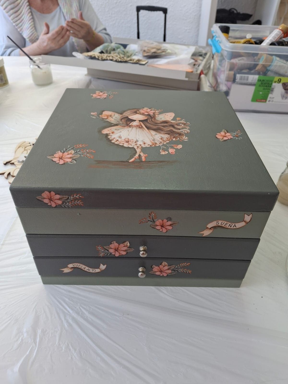Una caja de manualidades gris con varios cajones, decorada con una ilustración de hadas y detalles florales.