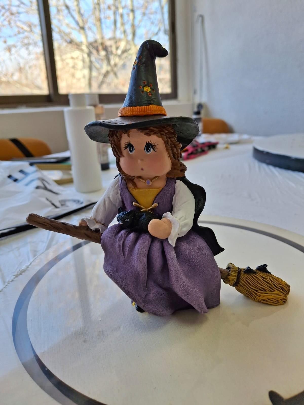 Una pequeña figurita de arcilla que representa a una bruja con un vestido morado y un sombrero negro montada en una escoba.