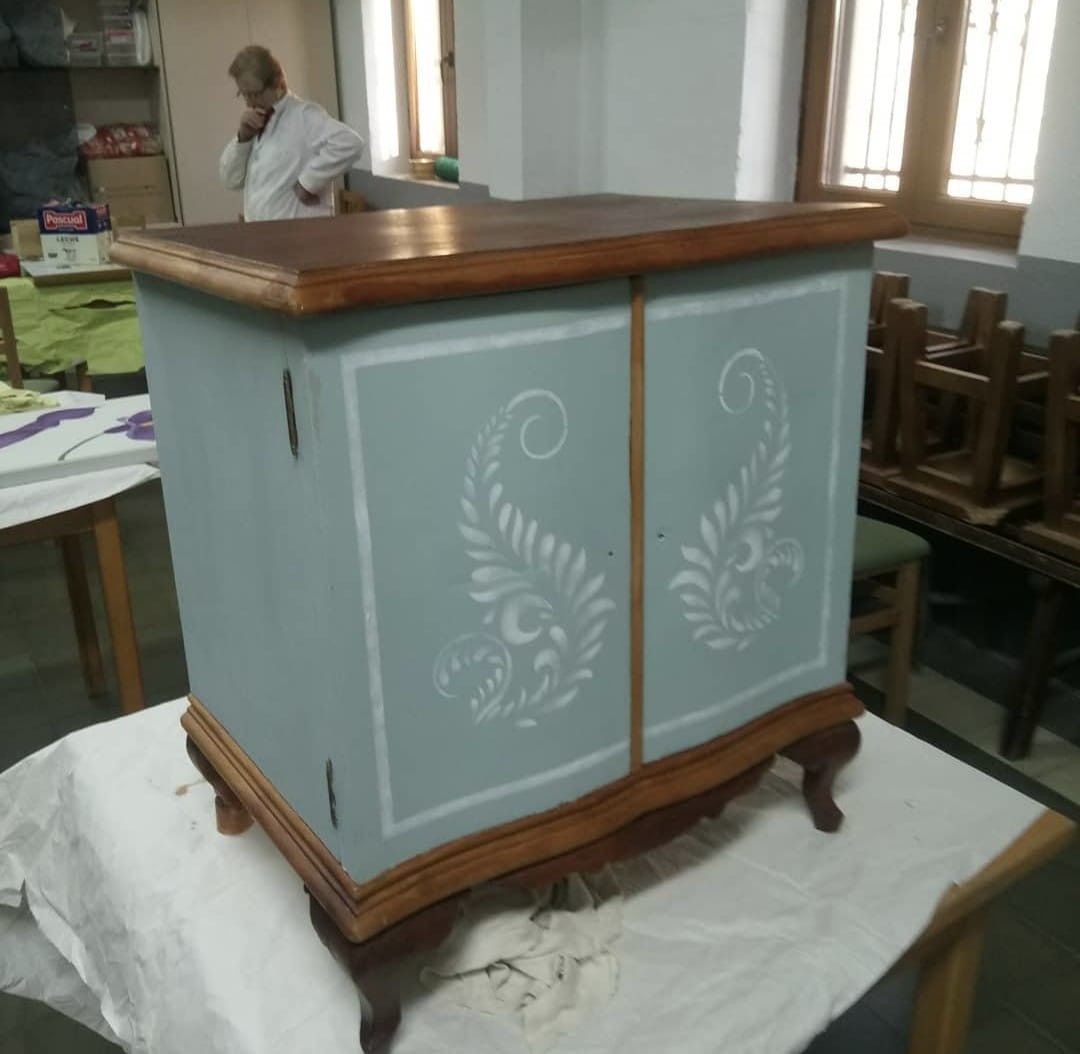 Un armario pintado de azul con diseños florales blancos decorativos en las puertas, que reposa sobre una mesa en un taller.
