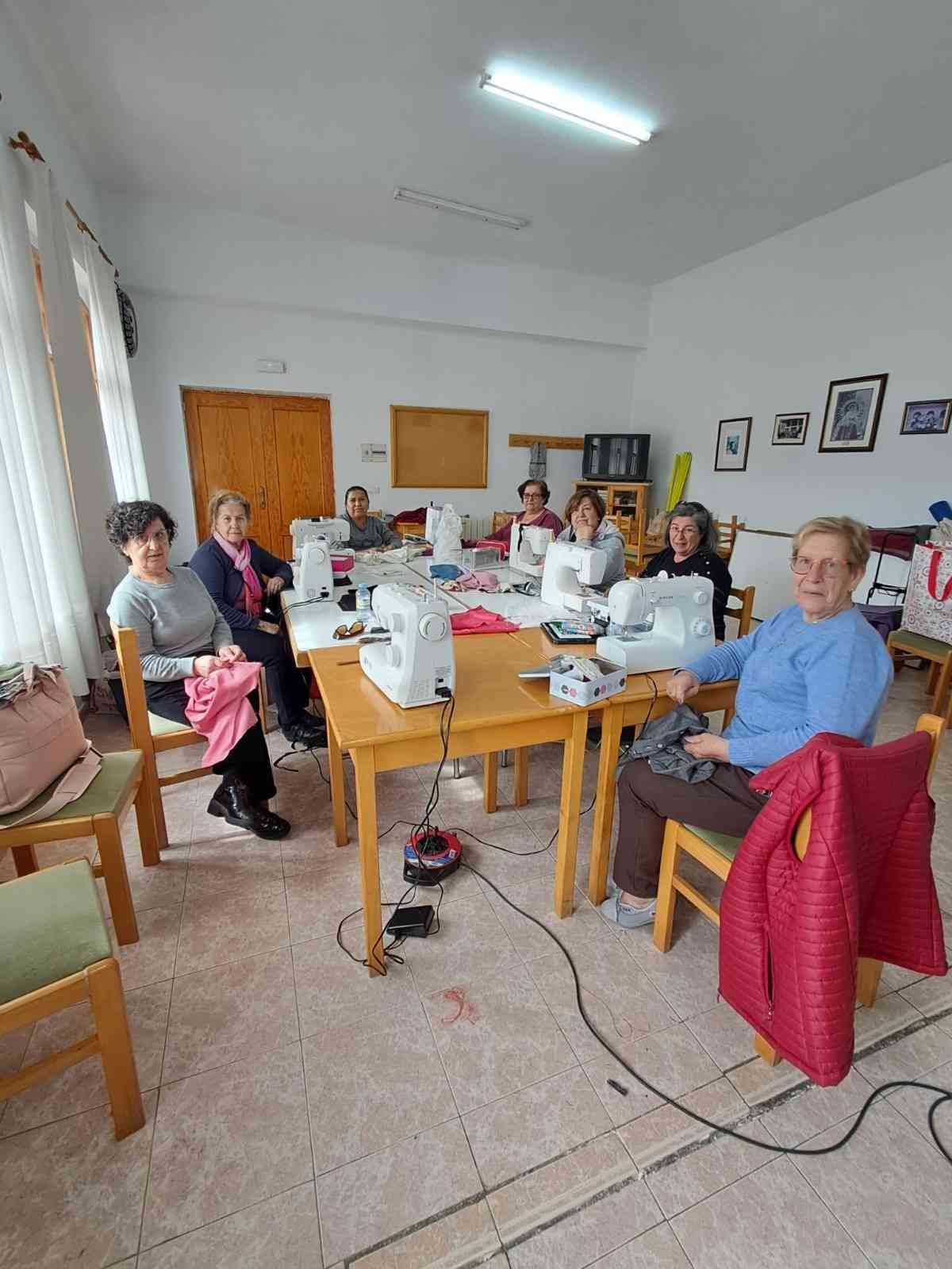 Un grupo de personas está sentado alrededor de mesas trabajando en proyectos de costura con máquinas en un aula luminosa.