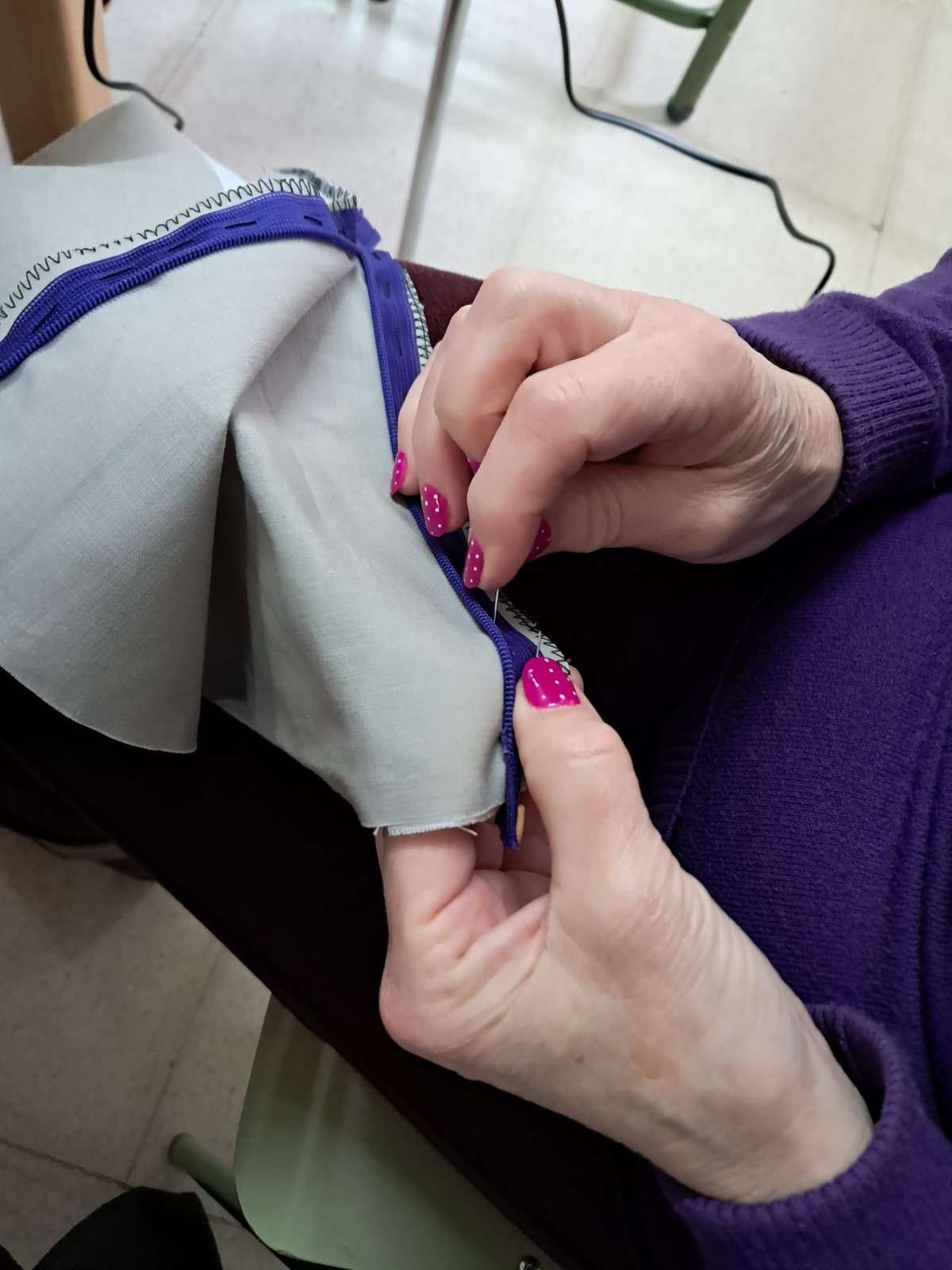 Unas manos con las uñas pintadas de rosa sostienen un trozo de tela gris claro con un ribete morado