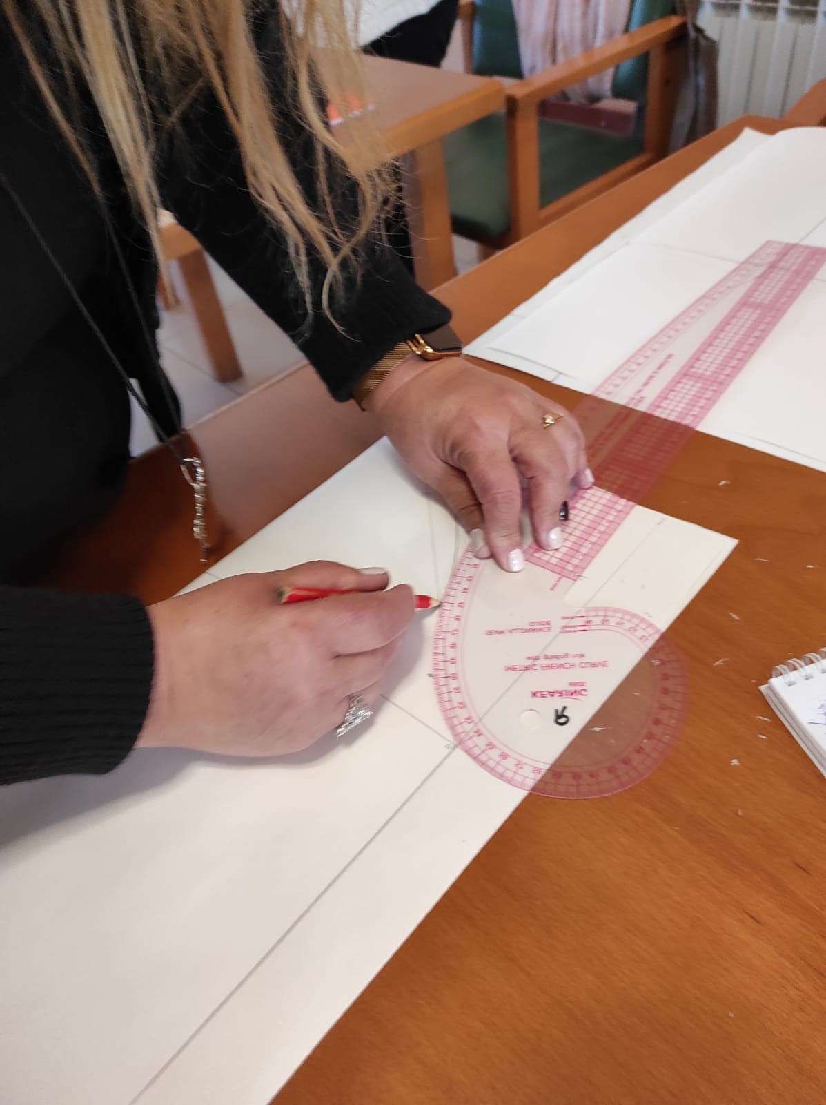 Sobre una mesa de madera, las manos trazan un patrón curvo sobre papel blanco utilizando un lápiz rojo 