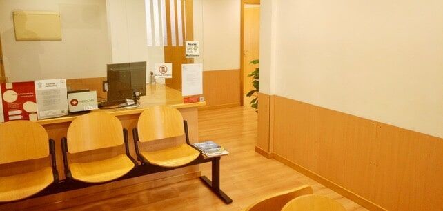 Área de recepção de um consultório médico com cadeiras de madeira, mesa e um corredor ao fundo.