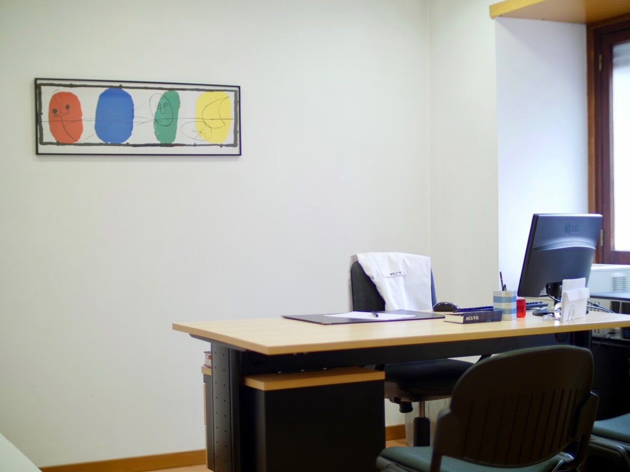 Interior de consultório médico com mesa de exame, escrivaninha, cadeiras e equipamentos médicos. Acabamento em madeira.