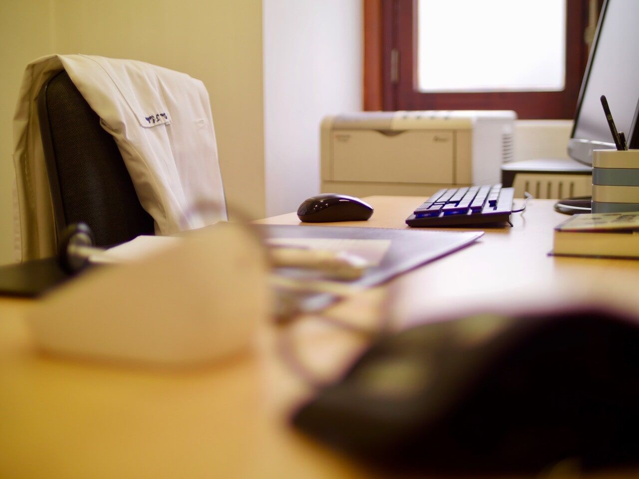 Mesa de escritório com computador, teclado, mouse e cadeira, além de jaleco.