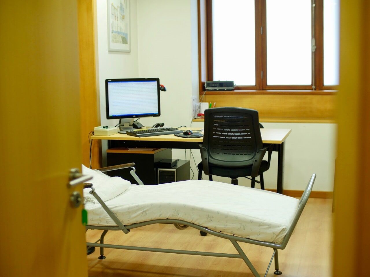 Sala de exame médico com cama, mesa com computador e cadeira. Janela iluminada ao fundo.