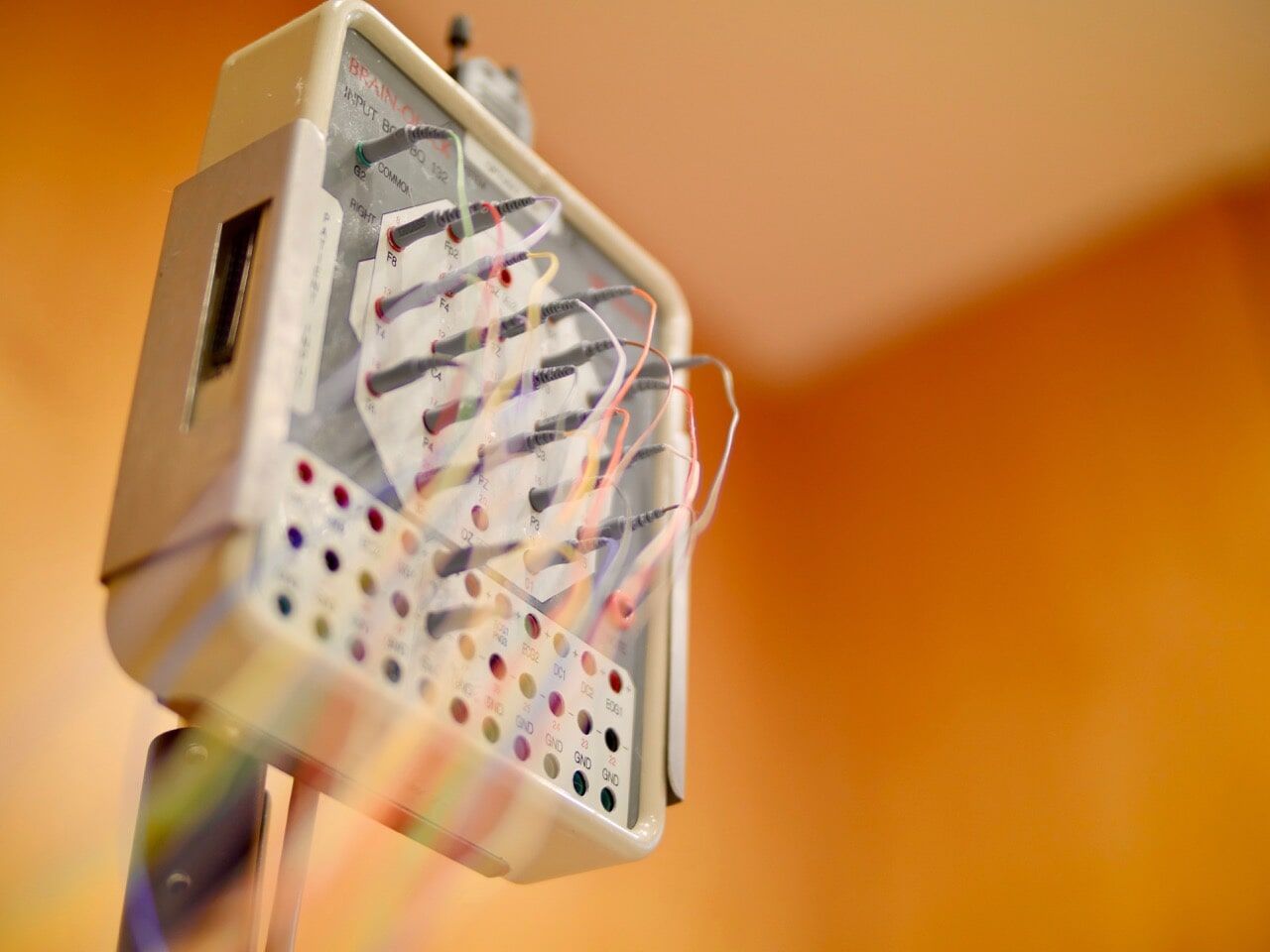Imagem em close-up de um aparelho de eletroterapia branco com várias entradas e fios coloridos, sobre um fundo laranja.