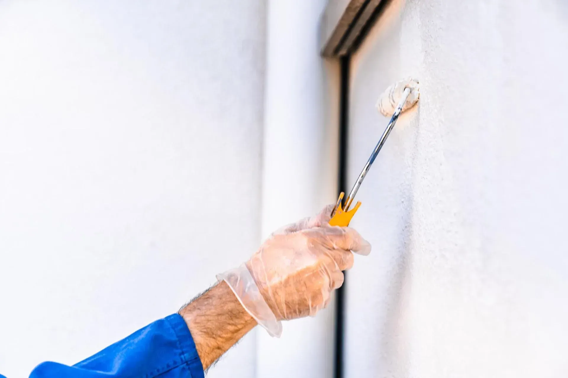 Una mano enguantada utiliza un rodillo de pintura para aplicar pintura blanca a una pared cerca del marco de una ventana.
