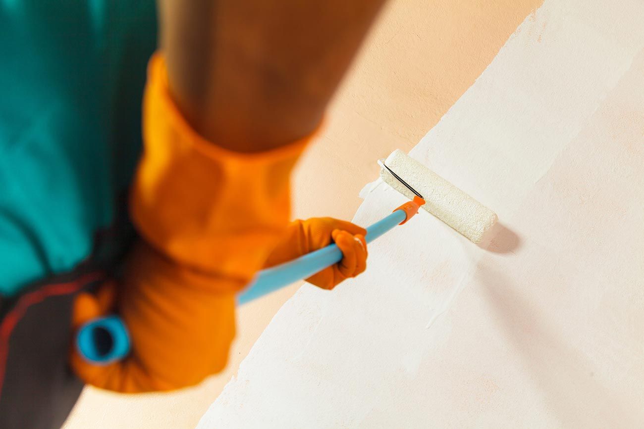 Persona pintando una pared blanca con un rodillo, con guantes naranjas y una manga azul.