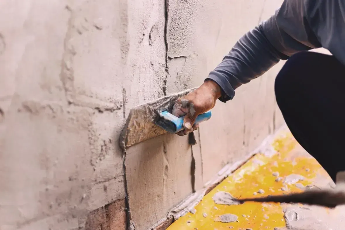 Un trabajador aplica yeso a una pared con una llana, con un revestimiento de suelo amarillo cerca.