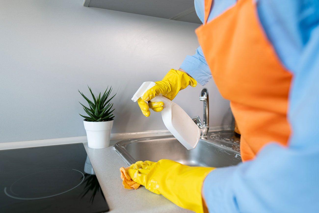 Una persona con guantes amarillos rocía con agua el fregadero de una cocina junto a una estufa y una maceta.