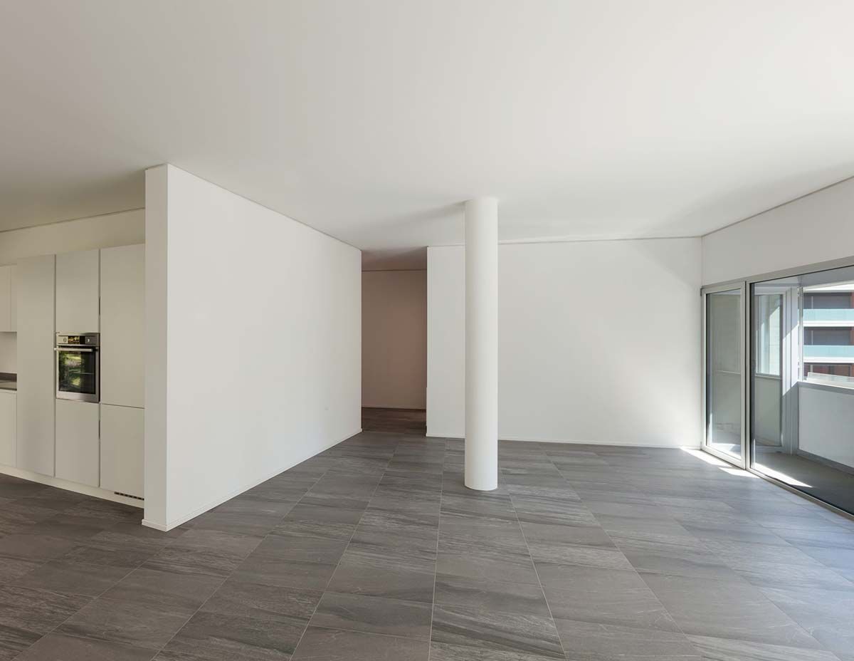 Sala de estar moderna y vacía con paredes blancas, suelo de baldosas grises, columna central y grandes ventanales.