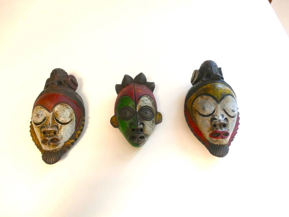 Trois masques sculptés colorés fixés au mur blanc, disposés en rangée.