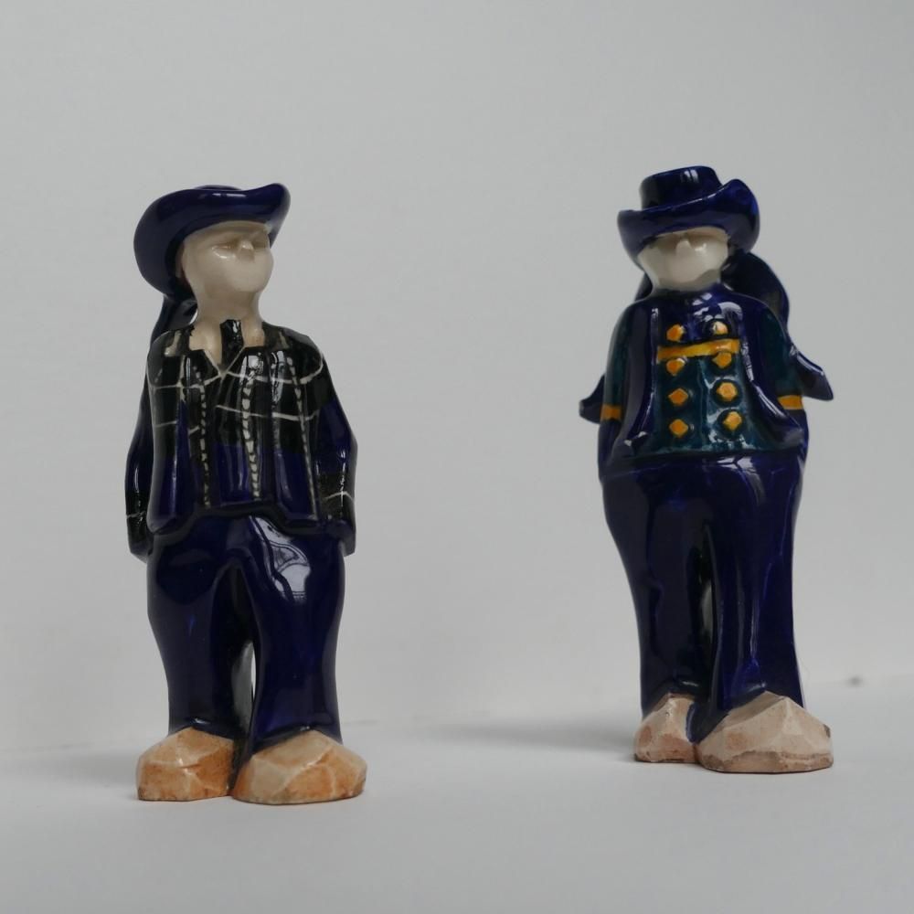 Deux petites figurines en céramique vêtues de bleu marine et coiffées de chapeaux, côte à côte sur un fond blanc uni