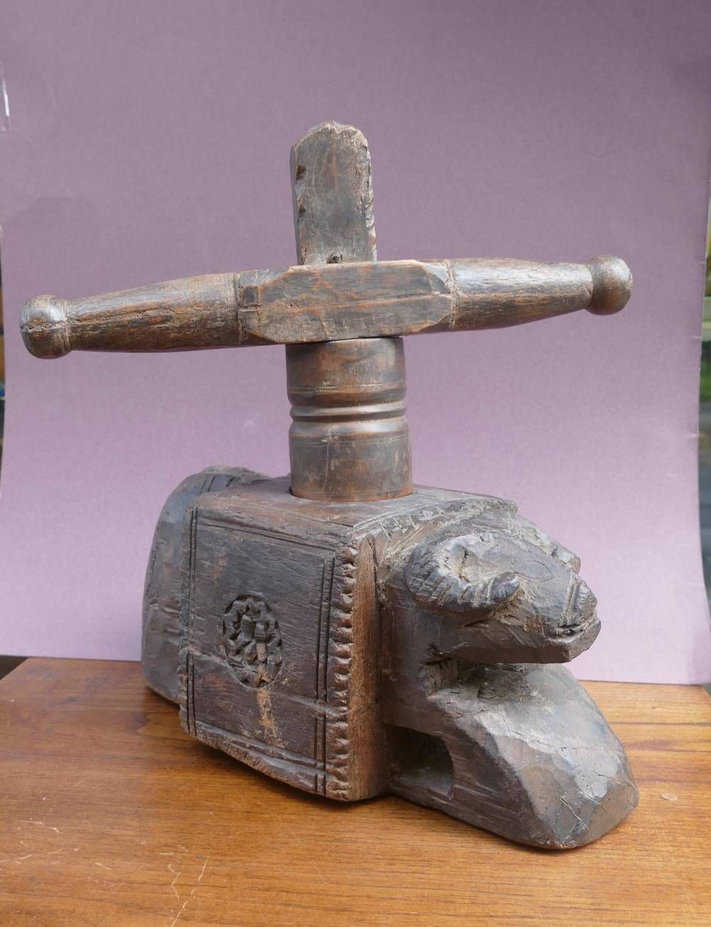 Broyeur ou pressoir rustique en bois sculpté, avec une poignée en T, posé sur une table en bois devant un mur violet