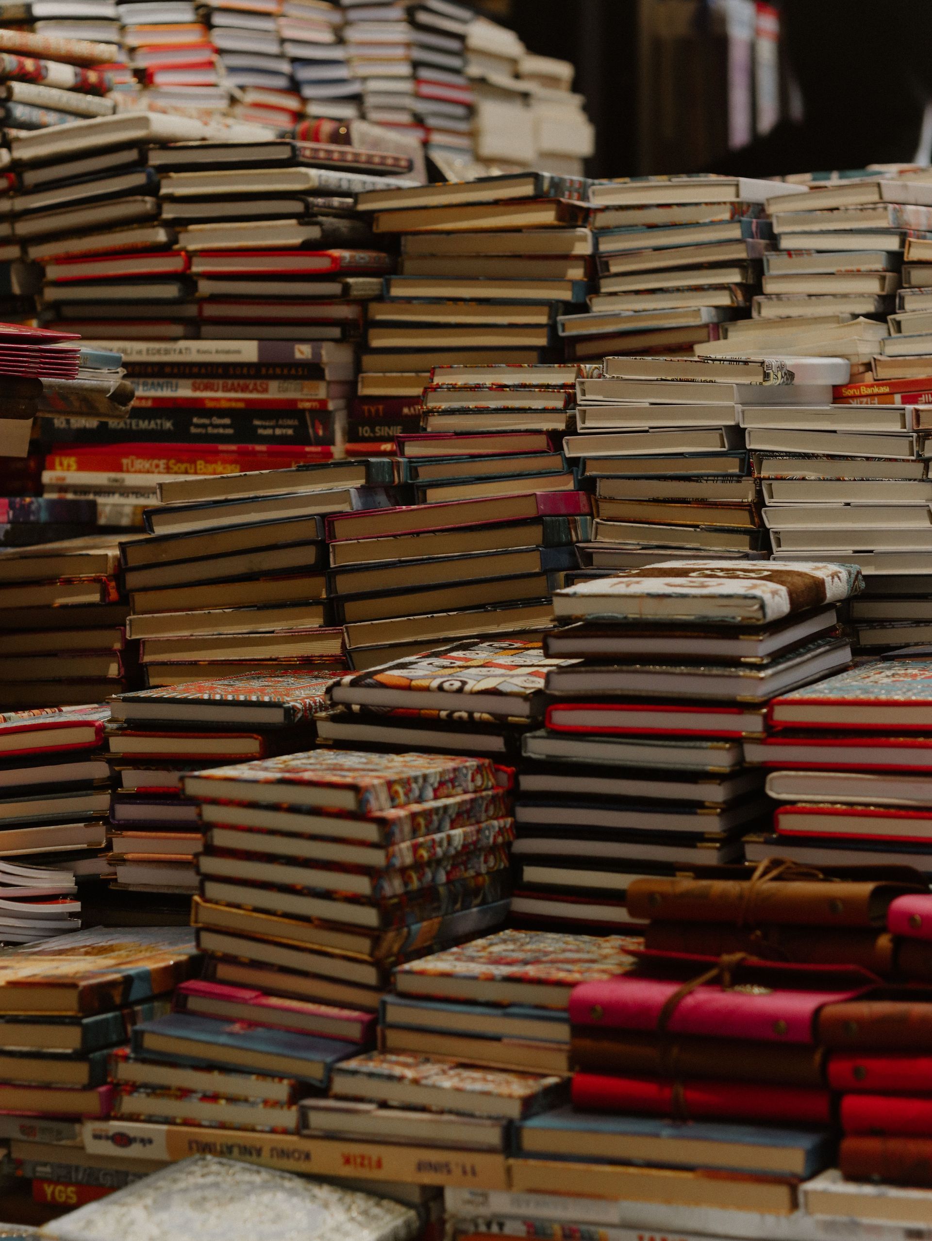 Des piles de livres variés, empilées très haut dans une librairie ou sur un étal de marché, créant une scène encombrée