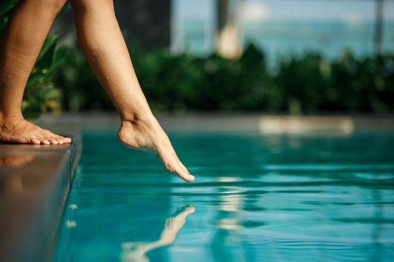 Eine Hand streckt sich nach einem türkisfarbenen Becken aus, die nackten Füsse stehen am Rand und die Hand spiegelt sich im Wasser.