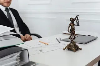Un abogado con traje está sentado en un escritorio. Sobre él hay una estatua de la Dama de la Justicia, papeles, una computadora portátil y un lápiz.