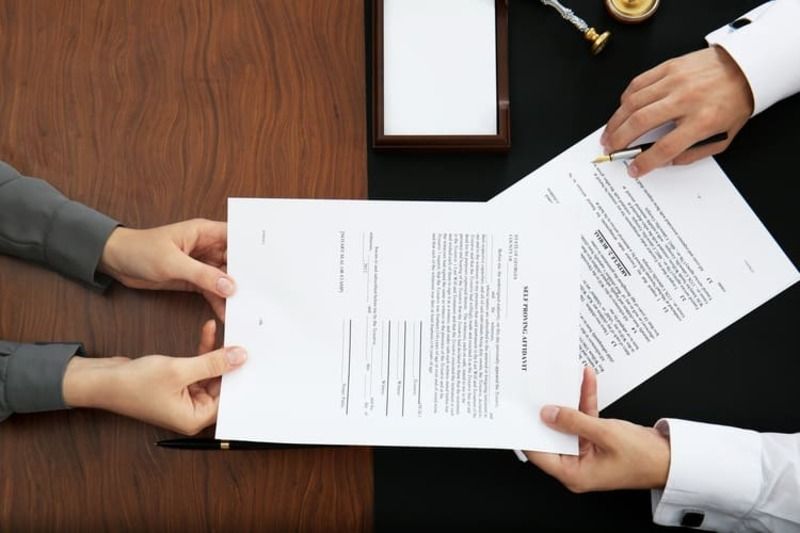 Dos personas intercambiando documentos sobre una mesa, posiblemente en un entorno legal o comercial.