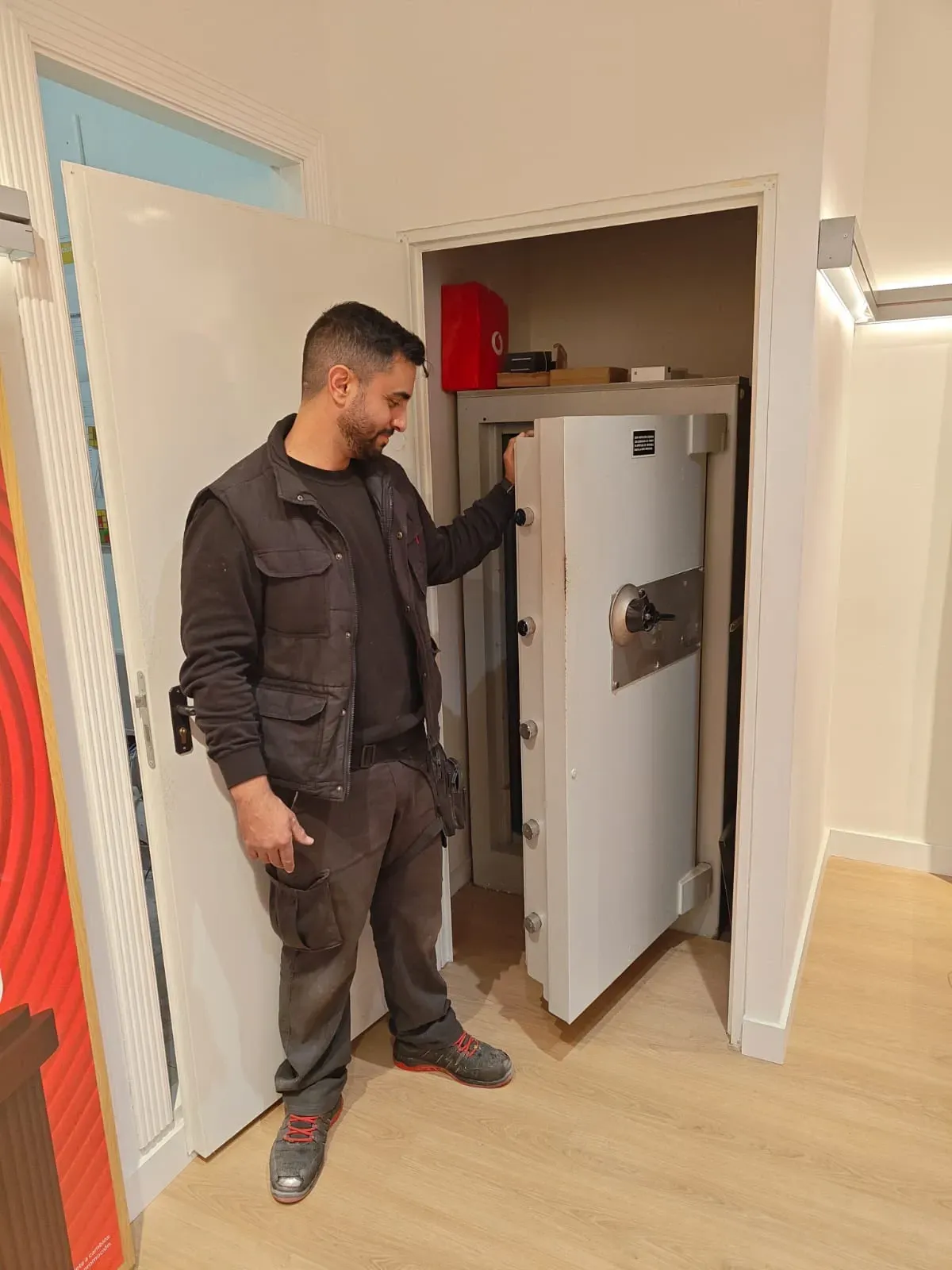 Hombre de pie junto a una gran caja fuerte abierta. Viste ropa de trabajo y la observa. Interior.