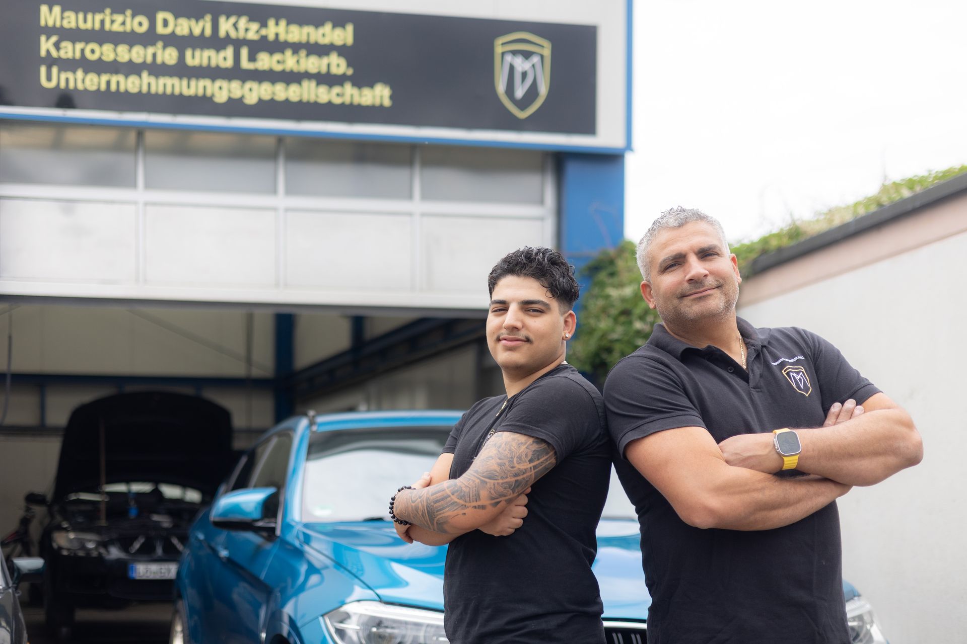 Zwei Männer stehen Rücken an Rücken vor einem blauen Auto.