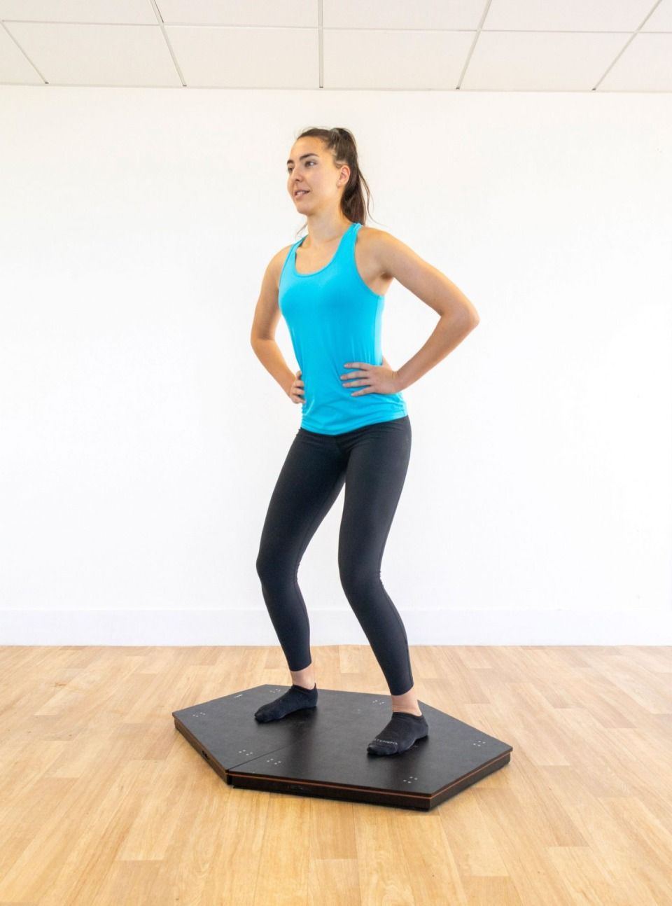 Una donna in abbigliamento sportivo si tiene in equilibrio su una balance board esagonale nera, con le mani sui fianchi, in palestra.