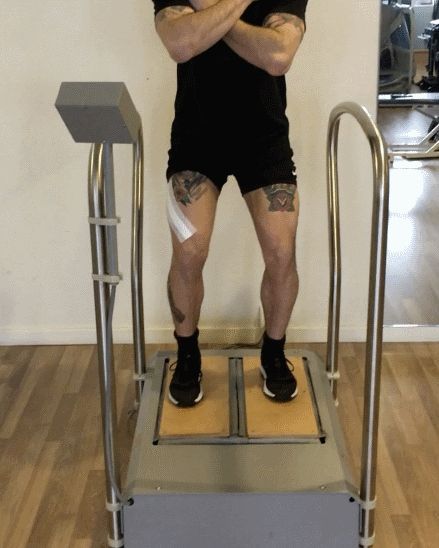 Una persona è in piedi su un dispositivo, con le braccia incrociate e del nastro adesivo sulla gamba, probabilmente in palestra.