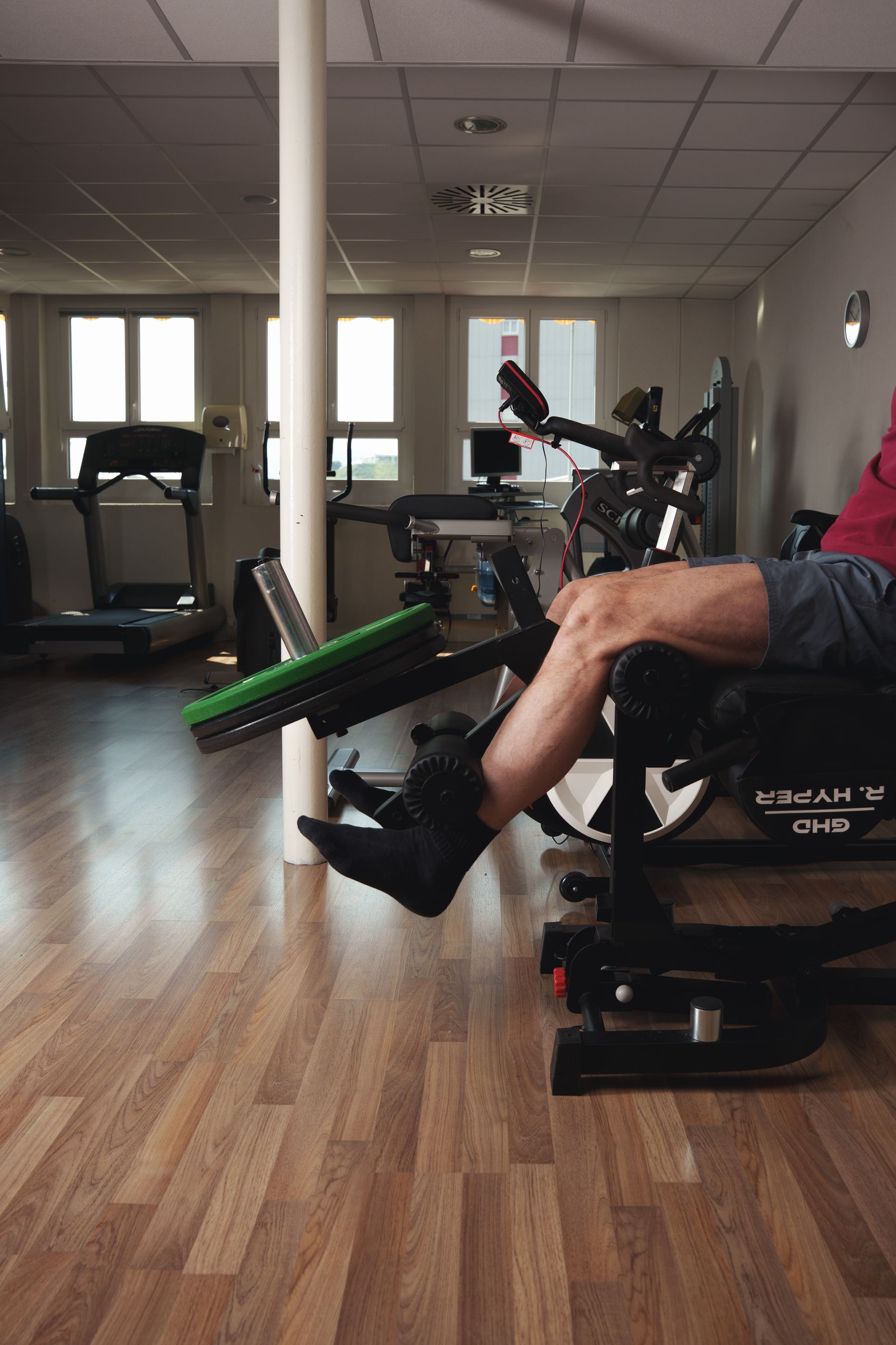 Uomo in piedi su una piattaforma, braccia incrociate, gambe visibili, con una benda su una coscia, in una palestra.
