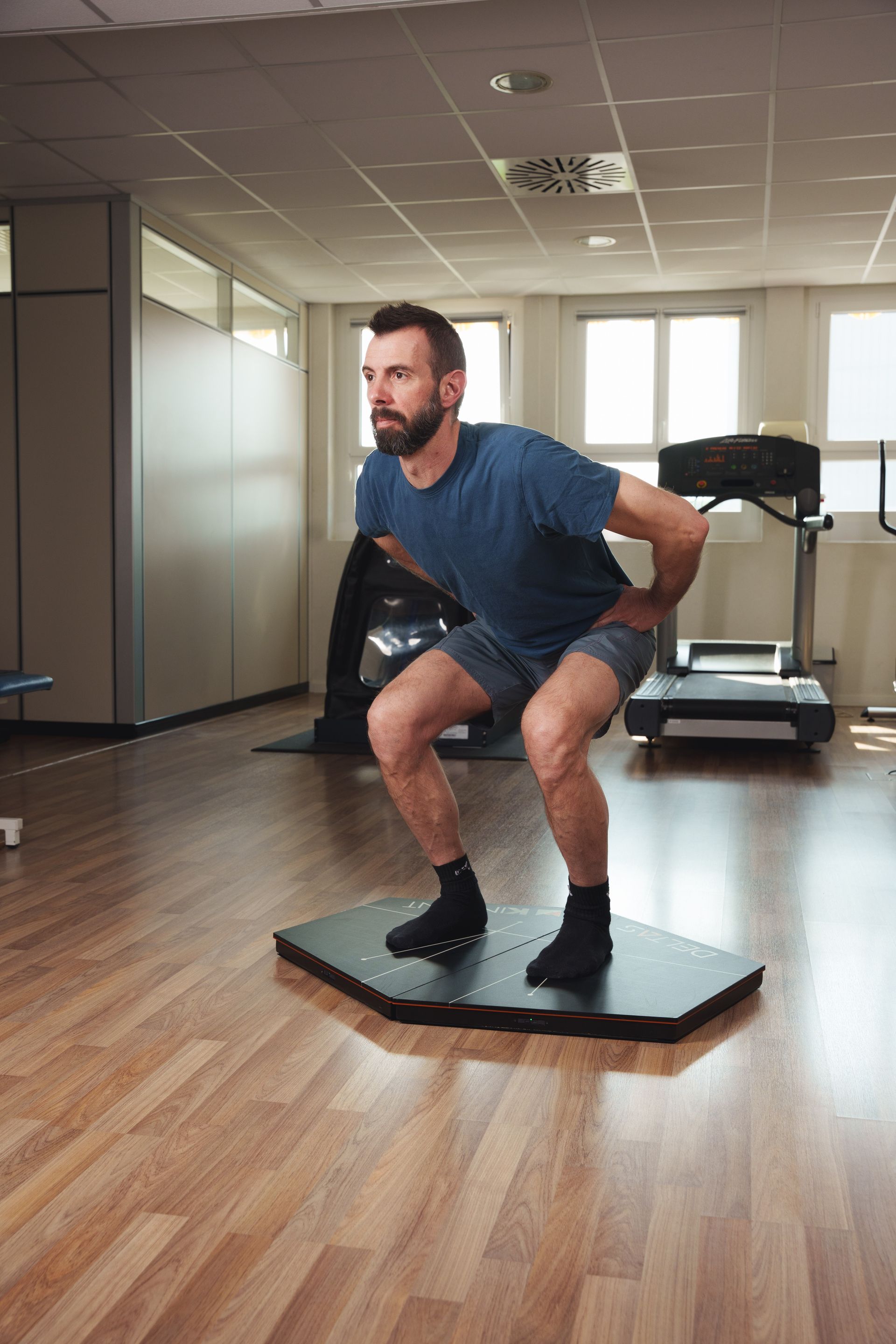 Una donna in abbigliamento sportivo si tiene in equilibrio su una balance board esagonale nera, con le mani sui fianchi, in palestra.
