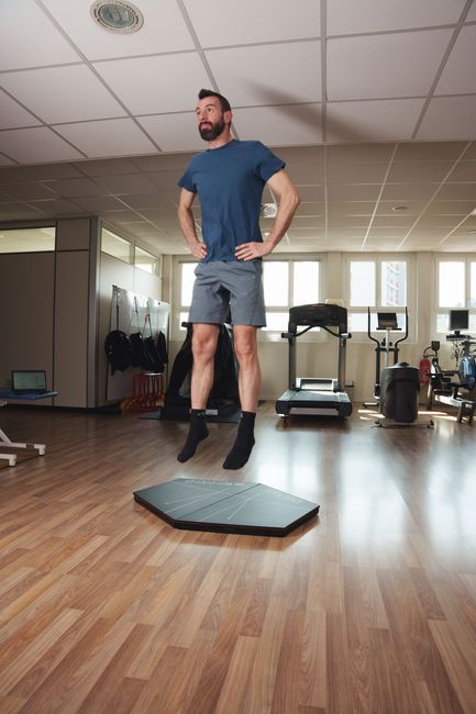 Una donna in abbigliamento sportivo si tiene in equilibrio su una balance board esagonale nera, con le mani sui fianchi, in palestra.