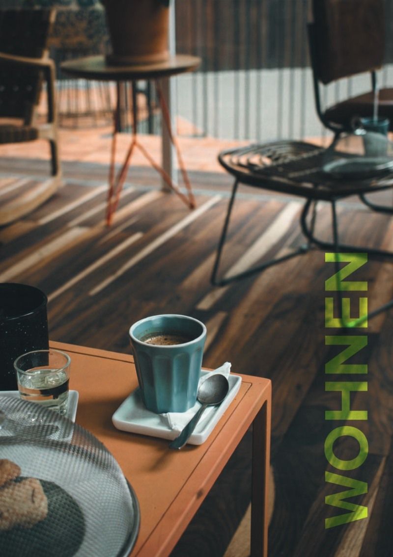 Innenaufnahme: Kaffeetasse auf Tisch mit „Wohnen“ in grüner Auflage auf dem Holzboden.