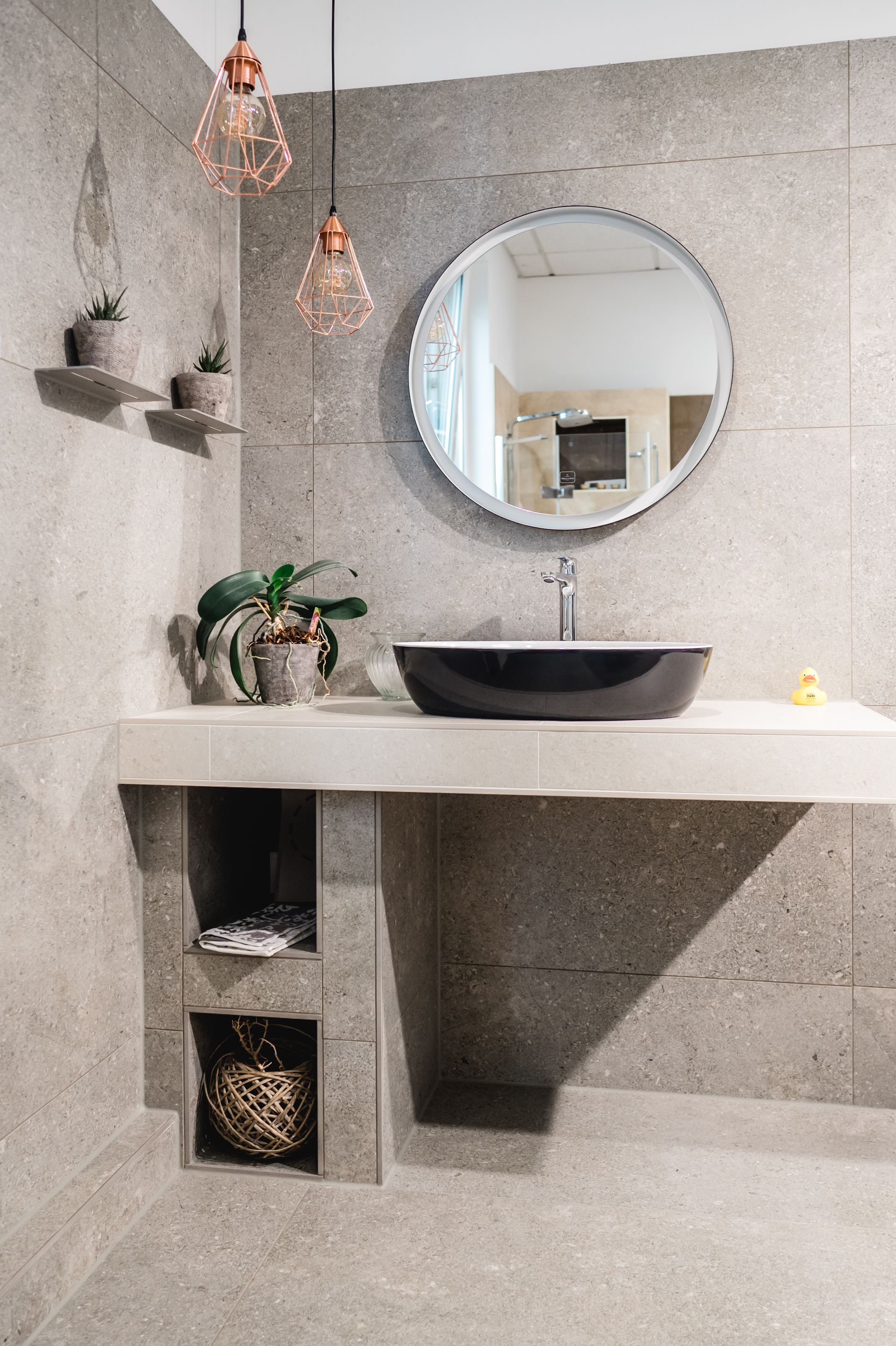 Modernes Badezimmer mit Betonwänden, schwebendem Regal, schwarzem Waschbecken, rundem Spiegel und Hängelampen.