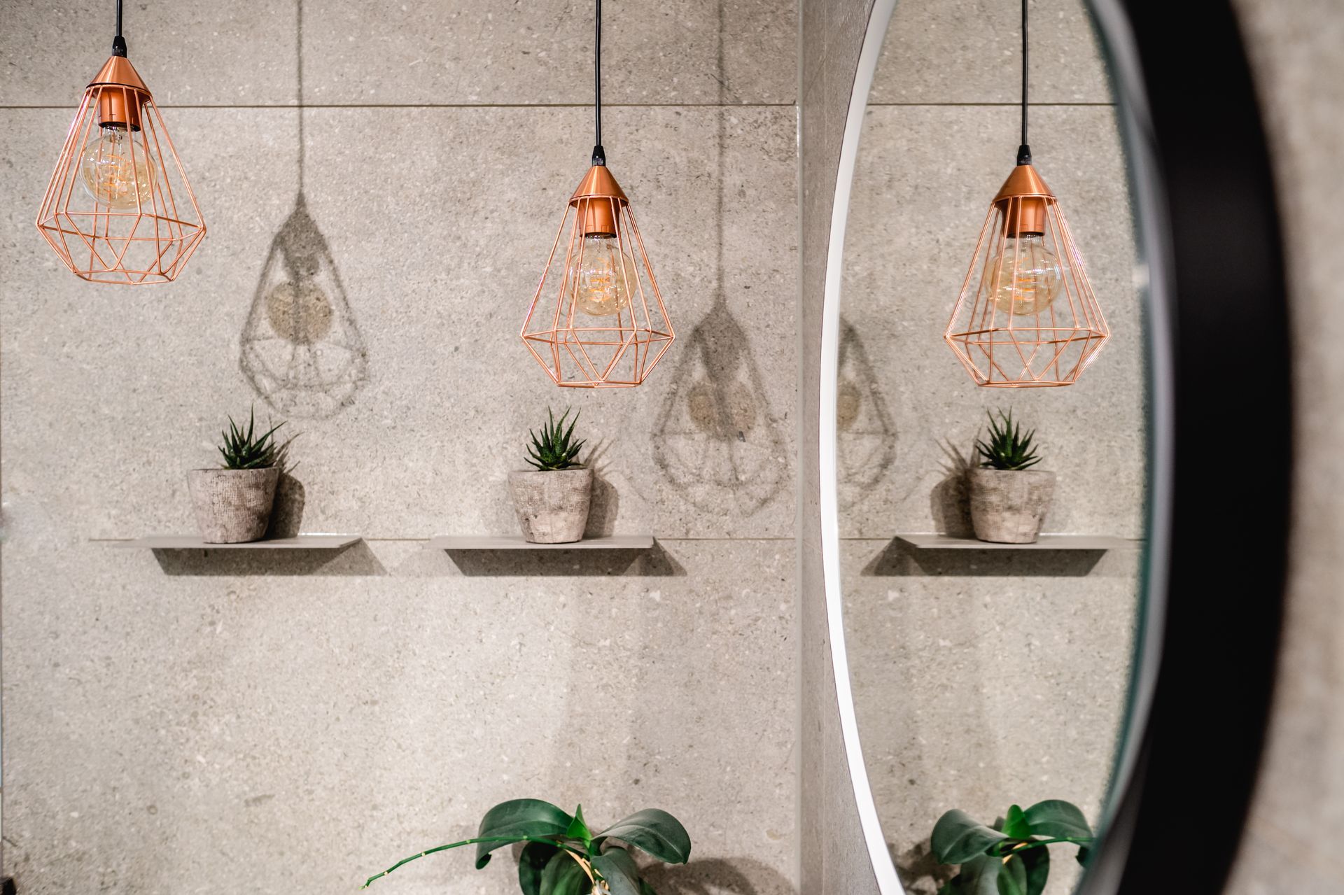 Kupferne Pendelleuchten werfen Schatten auf kleine Topfpflanzen auf Regalen. Ein runder Spiegel reflektiert die Szene.