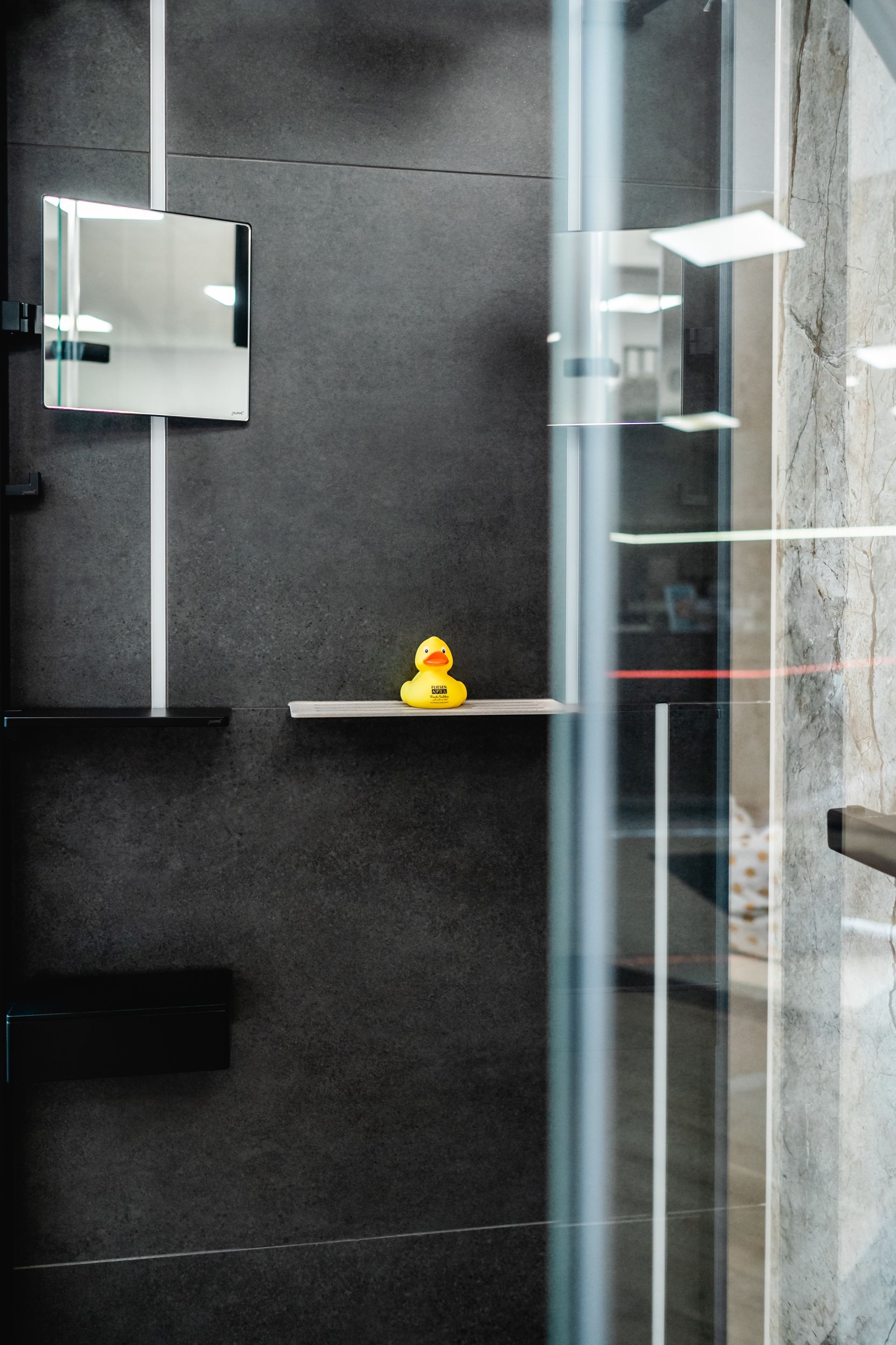Gelbe Gummiente auf einem grauen Regal in einer modernen Dusche mit Glastür.