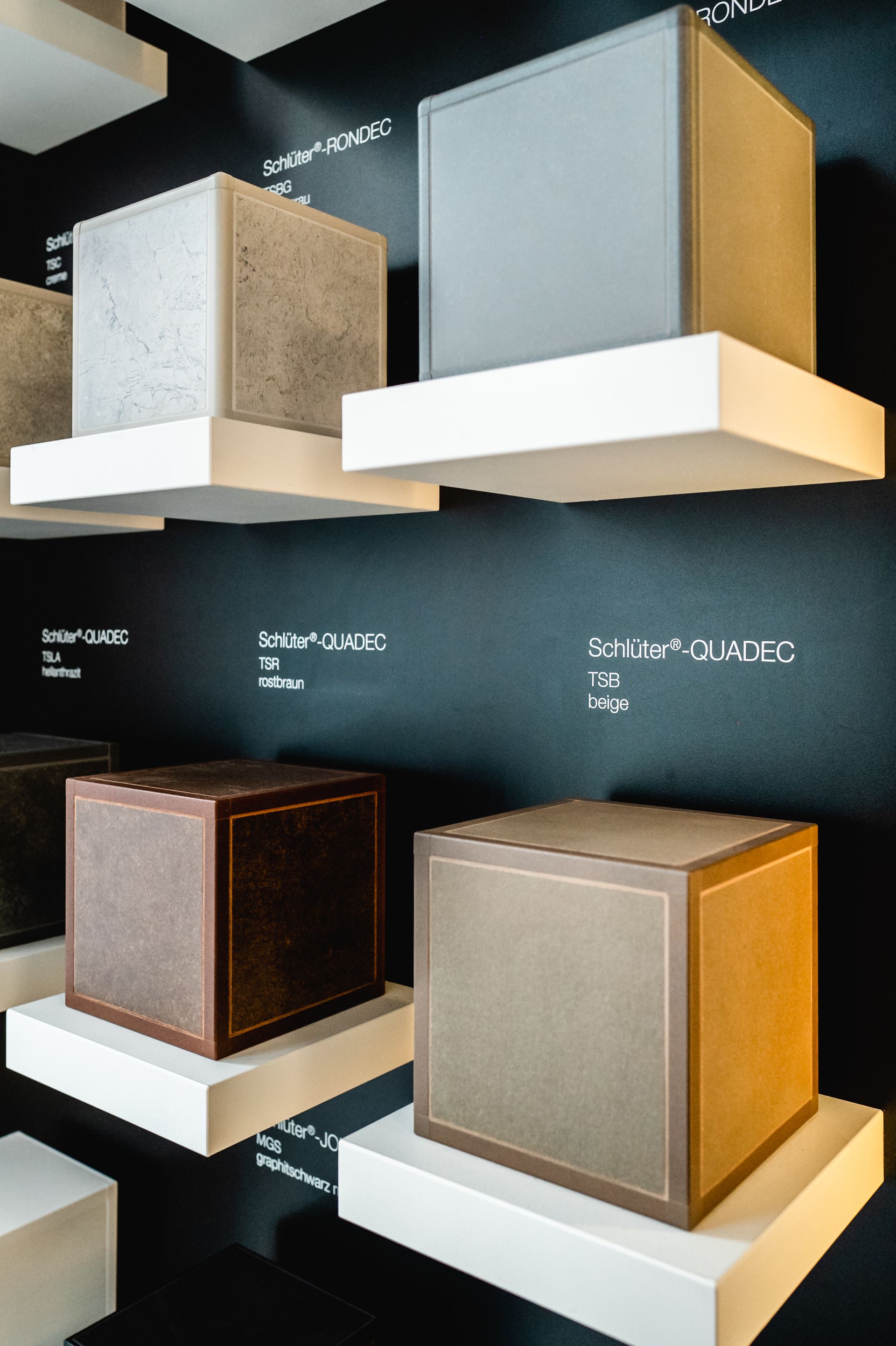 Würfel auf weißen Regalen vor einer schwarzen Wand, verschiedene Farben: Grau, Braun, Beige. Display für Produktmuster.
