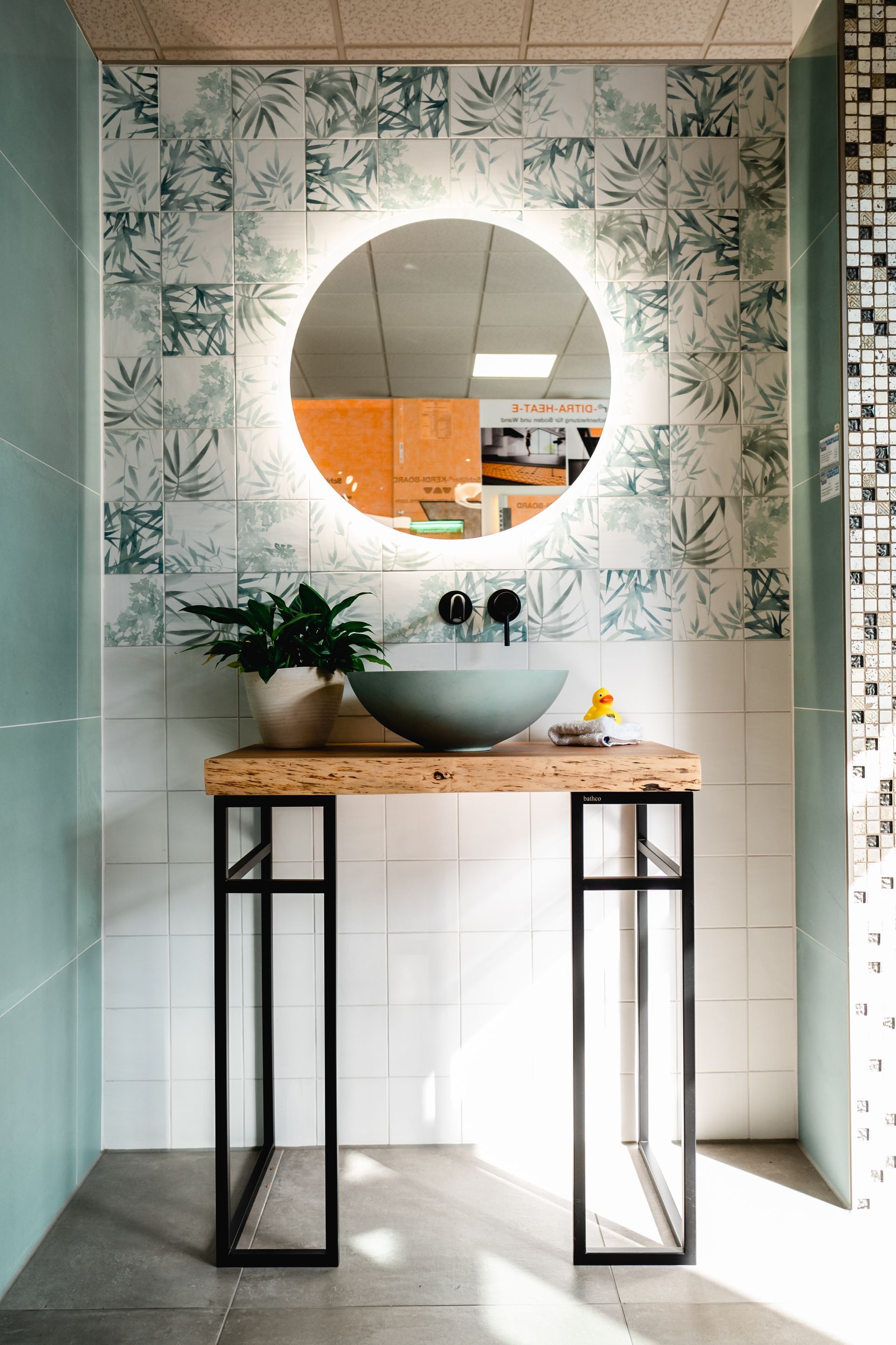 Badezimmer mit rundem, hinterleuchtetem Spiegel, blauem Aufsatzwaschbecken und Holztisch mit schwarzen Beinen vor gemusterten Fliesen.