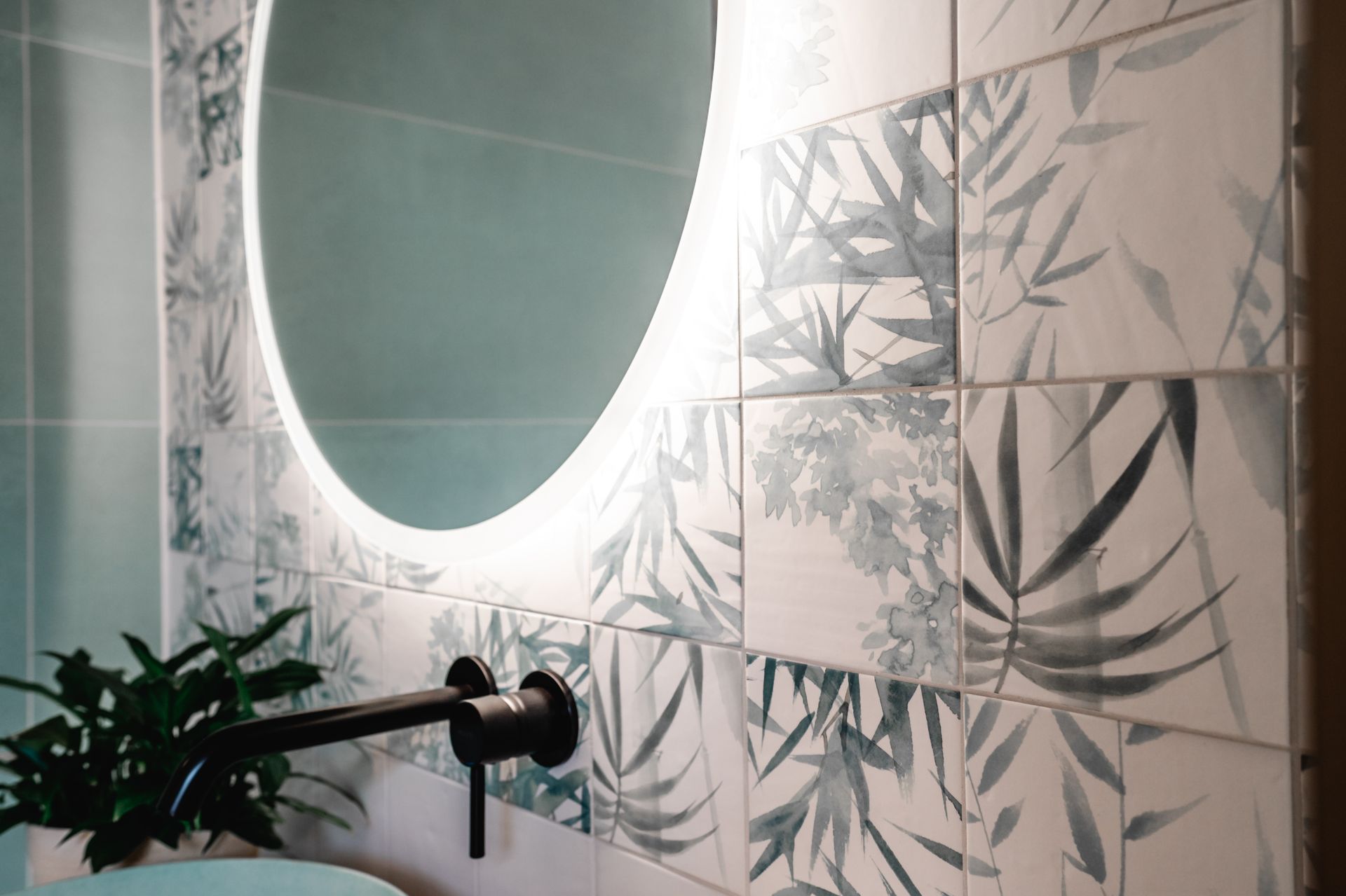 Ovaler Spiegel mit Hintergrundbeleuchtung über einem Waschbecken mit schwarzem Wasserhahn. Fliesenrückwand im Botanischen Stil, grüne Wand.
