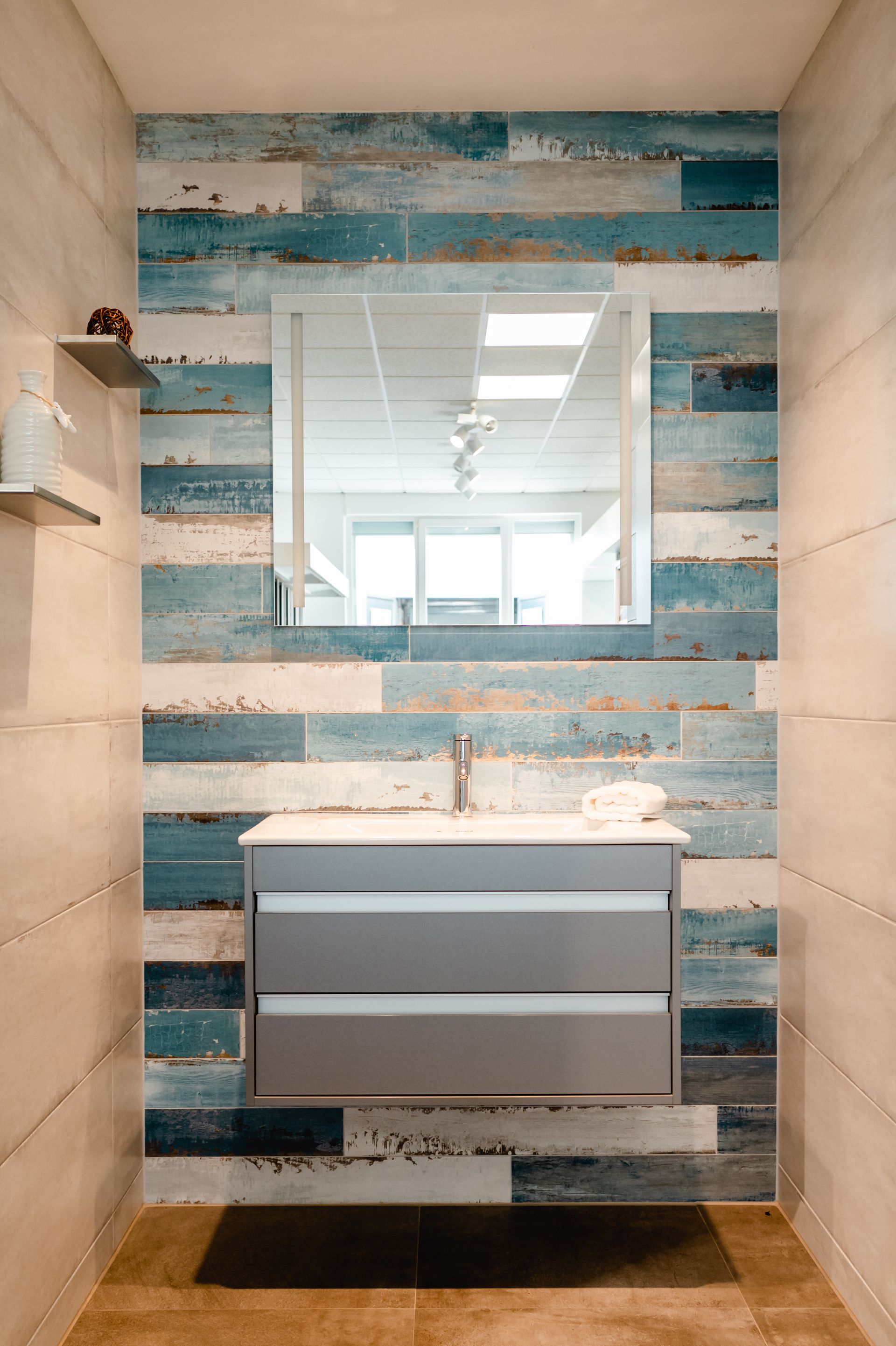 Badezimmer mit blau-weißer Akzentwand in Holzoptik, silbernem Waschtisch, quadratischem Spiegel und Regal.