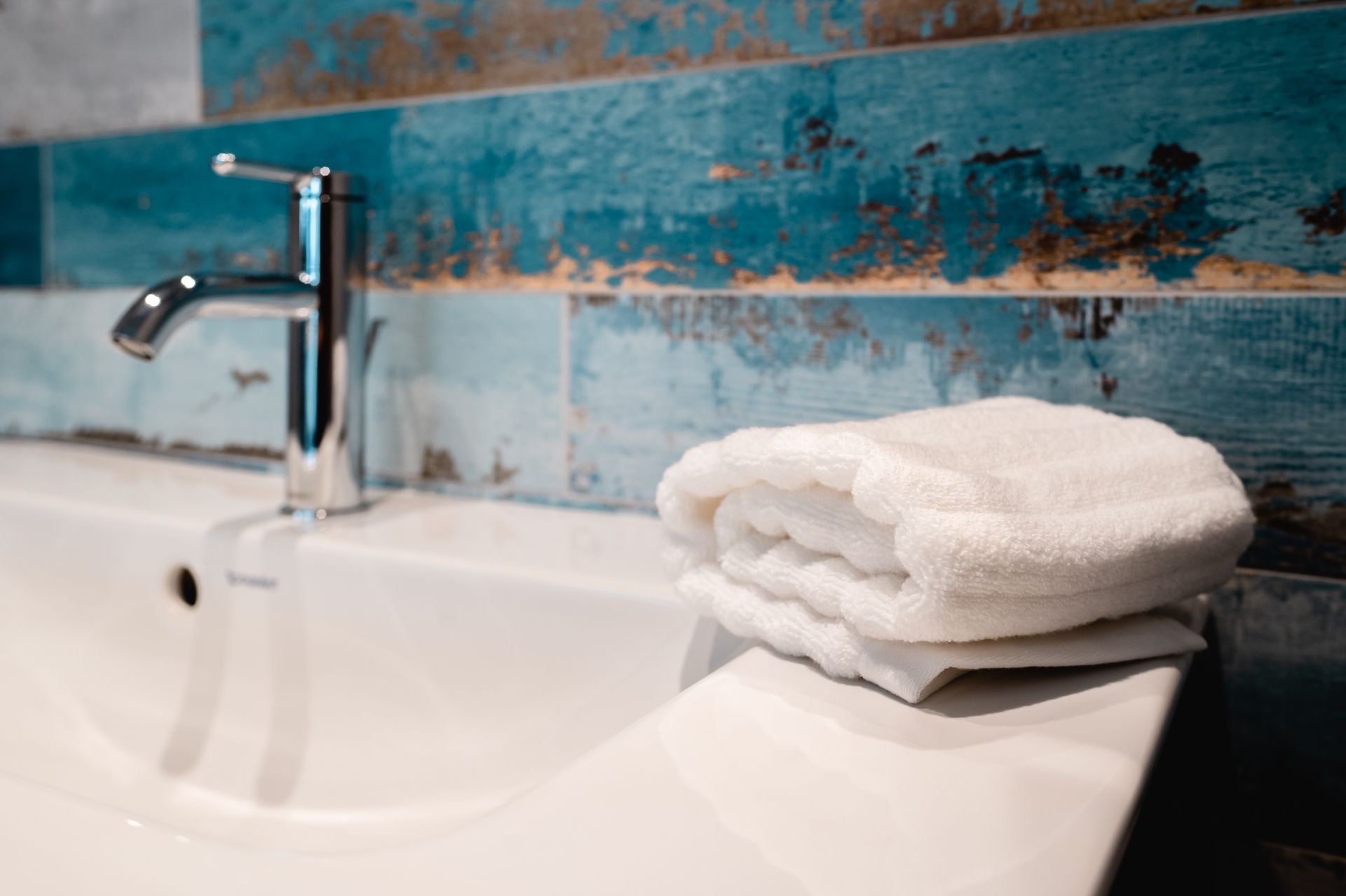 Weißes Handtuch auf einem weißen Waschbecken neben einem Chromhahn; blaugrüne und braune geflieste Wand im Hintergrund.