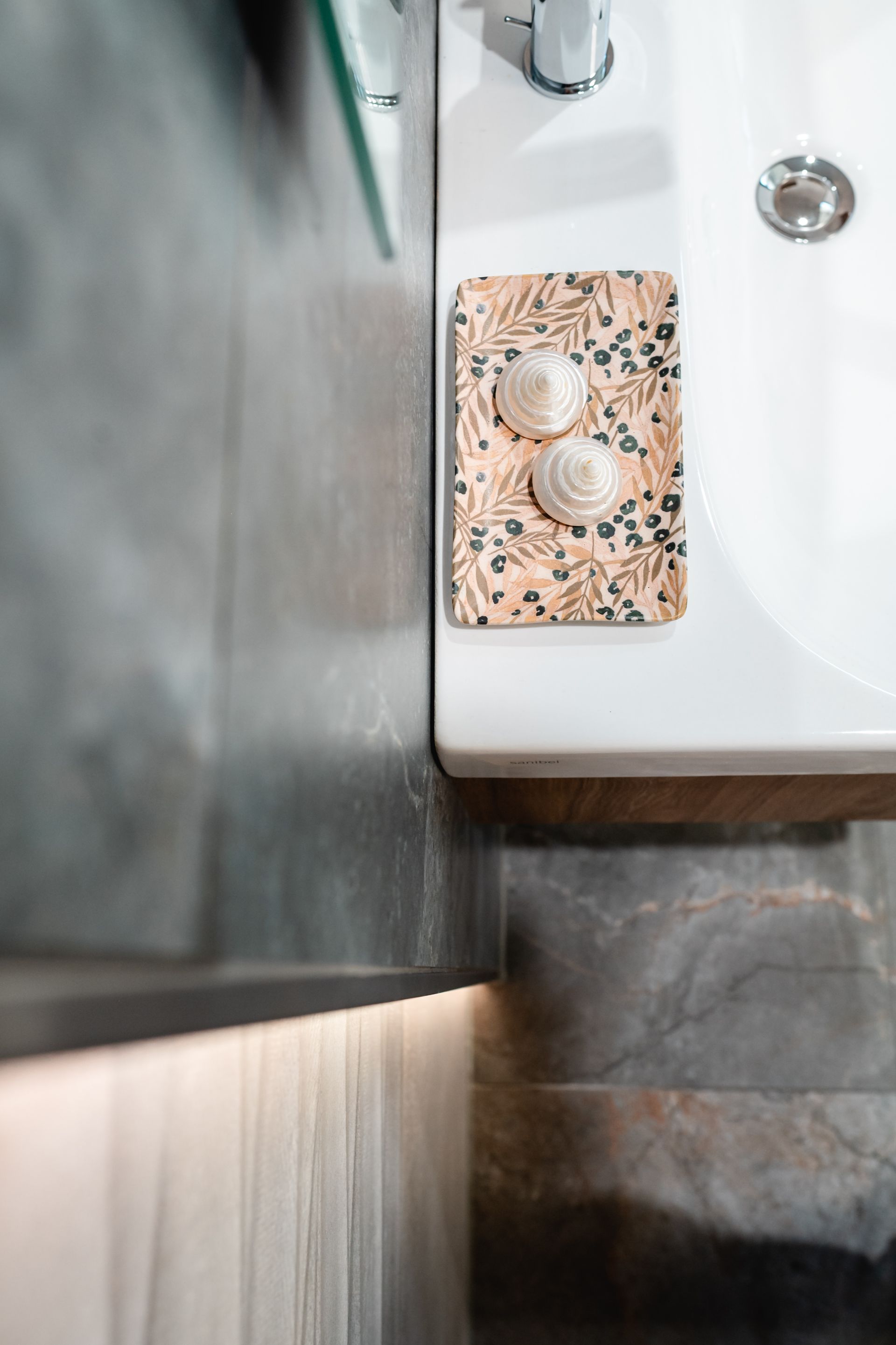 Zwei Muscheln auf einem gemusterten Tablett neben einem weißen Waschbecken. Graue Wand- und Unterschrankbeleuchtung.