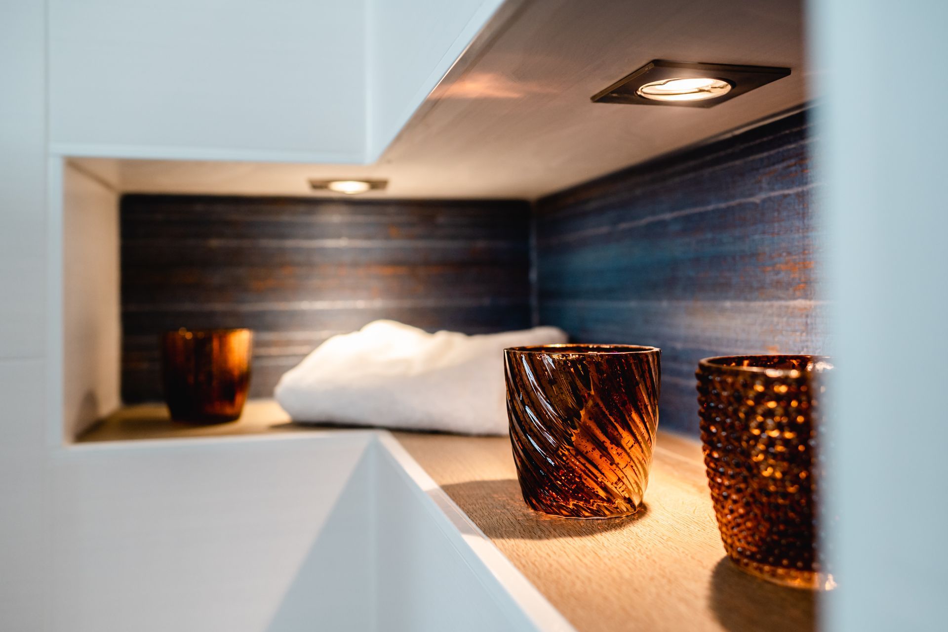 Eingelassenes Regal mit Votivkerzen aus Bernsteinglas, weißem Handtuch und einer blaubraunen, strukturierten Rückwand, beleuchtet durch Deckenstrahler.