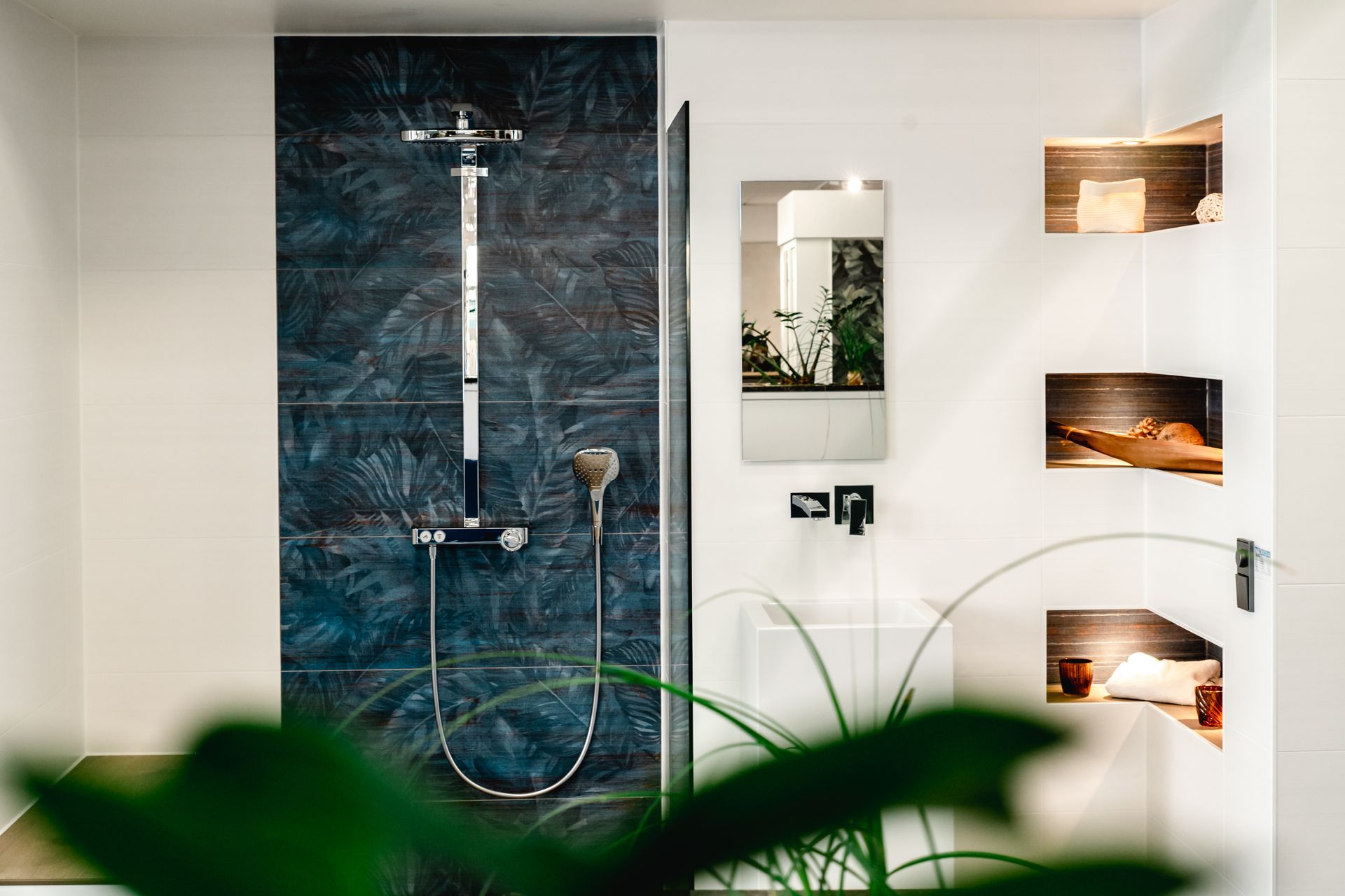 Dusche mit dunkelblauem Fliesenhintergrund, silbernen Armaturen und Regalen mit Gegenständen an einer weißen Wand.