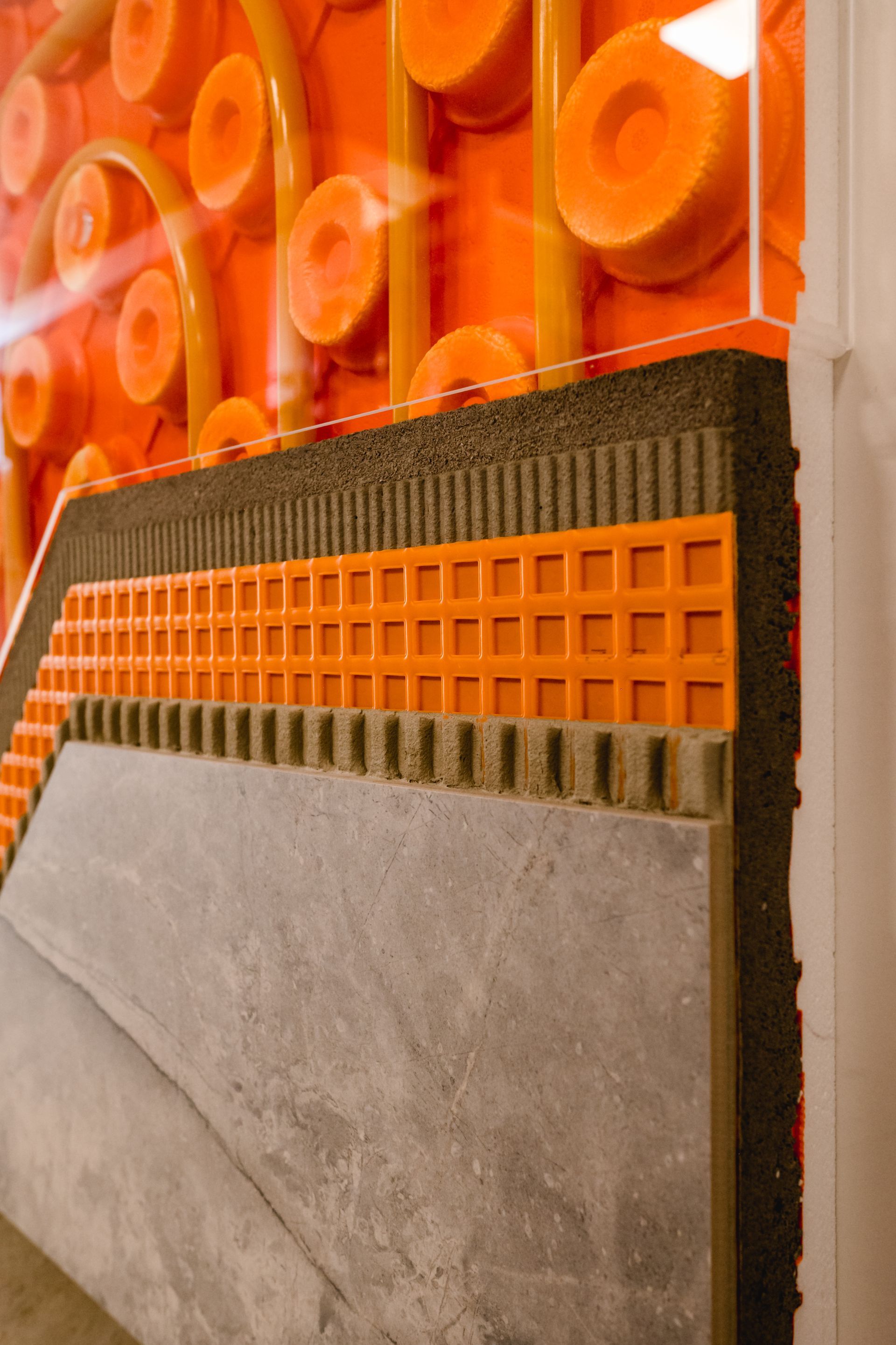 Schnittansicht einer Fußbodenheizung mit Betonschichten, Isolierung, orangefarbenen Rohren und Gitter.