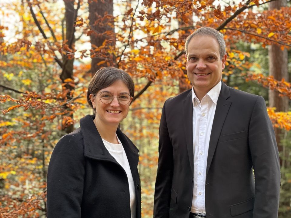 Frau und Mann lächeln vor herbstlichem Laub. Der Mann trägt ein Sakko, die Frau einen Mantel und eine Brille.