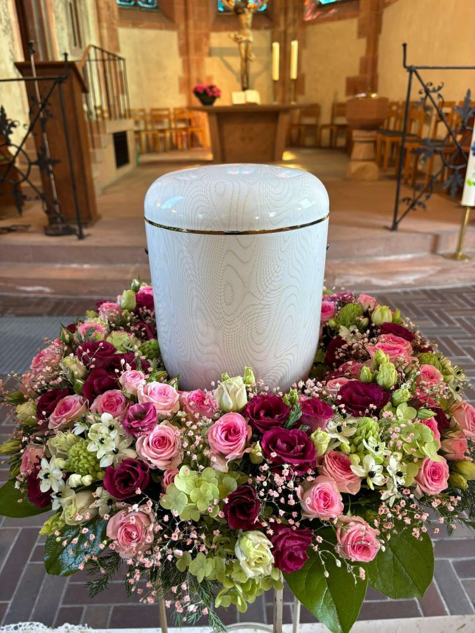 Vorbereitungen für eine Trauerfeier mit Kerzen, Blumengestecken, einem Kreuz und einer Urne.