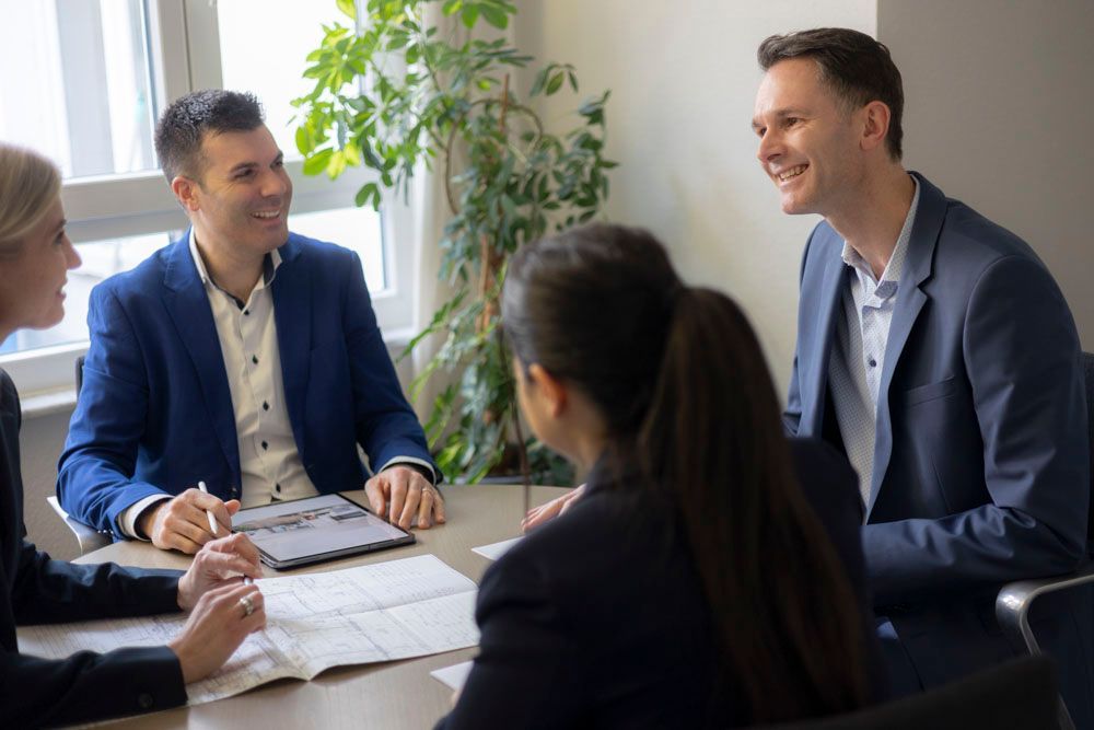Vier Geschäftsleute in Businesskleidung sitzen lächelnd in einem hellen Büro an einem Besprechungstisch und arbeiten mit Dokumenten zusammen.