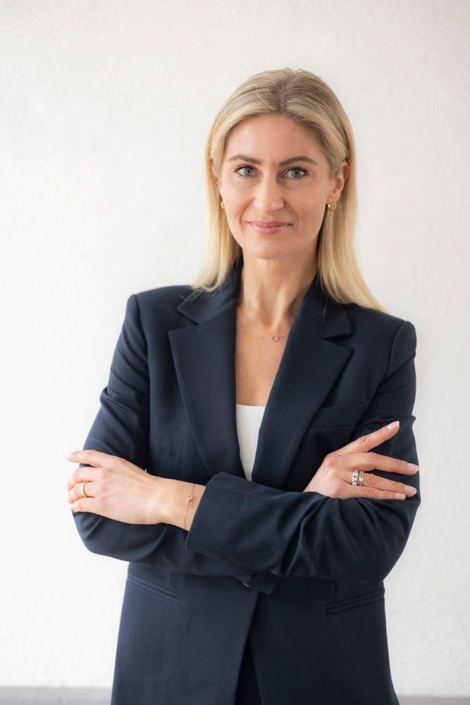 Eine Person mit blonden Haaren, die einen dunkelblauen Blazer trägt, steht mit verschränkten Armen vor einem weißen Hintergrund.