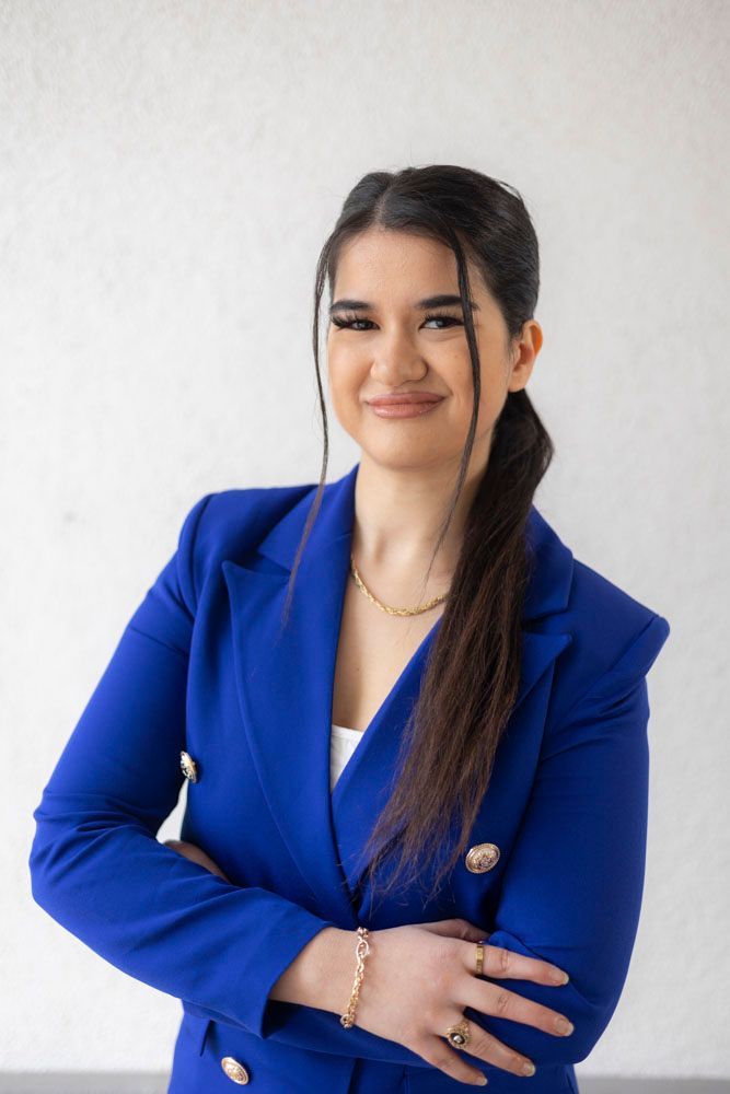 Eine Person mit dunklem, zu einem seitlichen Pferdeschwanz frisiertem Haar posiert mit verschränkten Armen und trägt einen hellblauen Blazer.