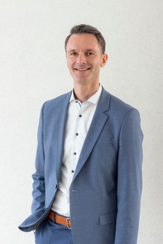 Lächelnder Mann im Anzug mit Krawatte vor weißem Hintergrund.