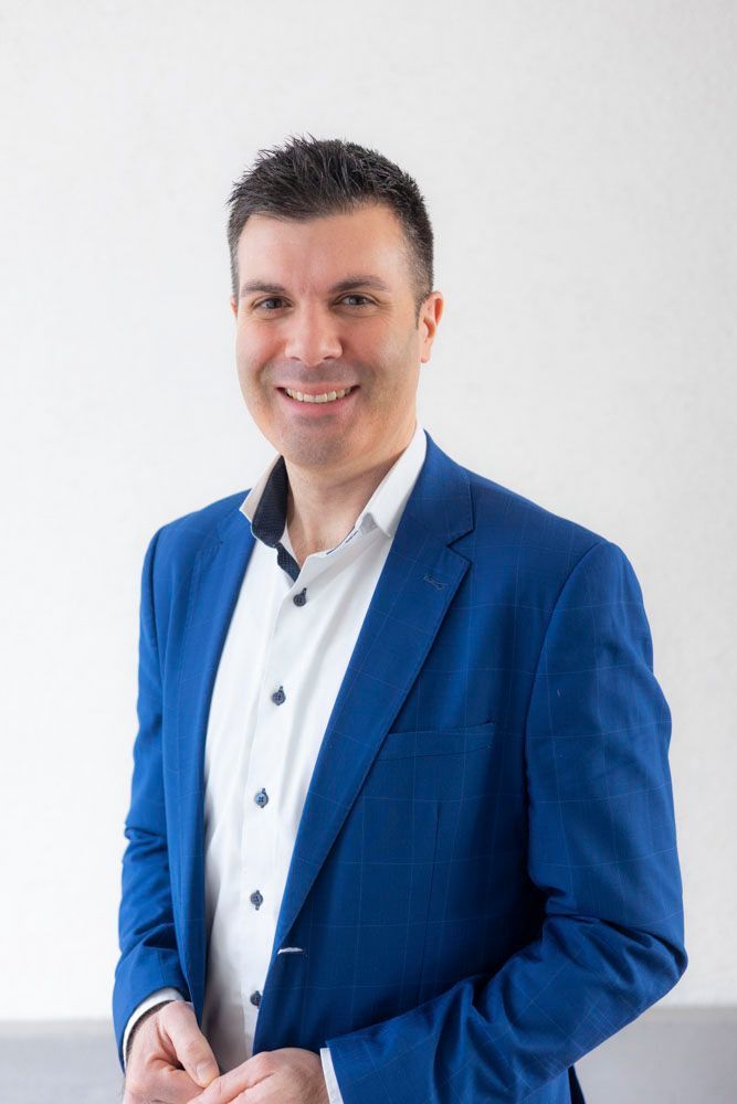 Eine lächelnde Person in einem blauen Blazer und einem weißen Hemd mit Knöpfen steht vor einem schlichten weißen Hintergrund.